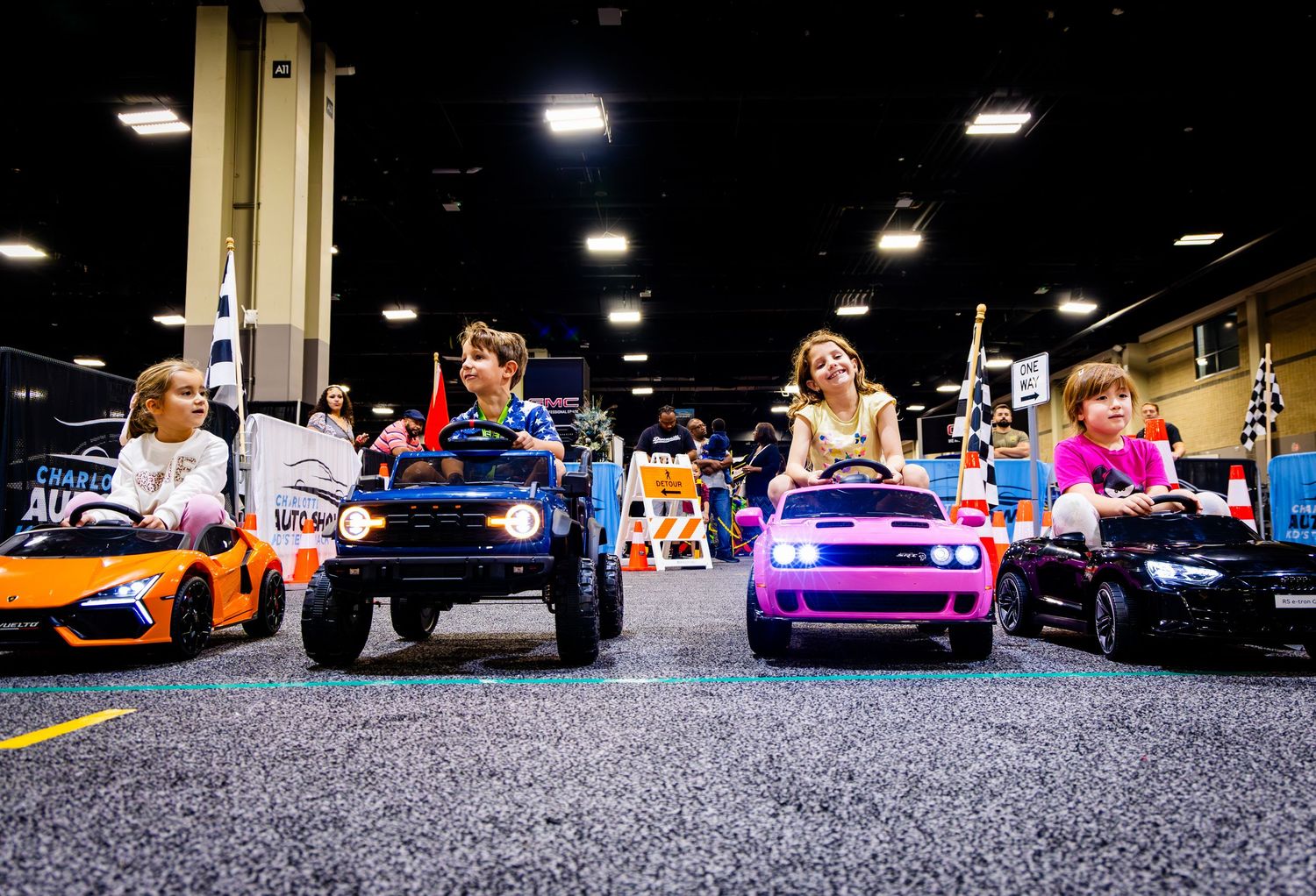 Colorful toy cars lined up on a carpeted floor at an indoor event.