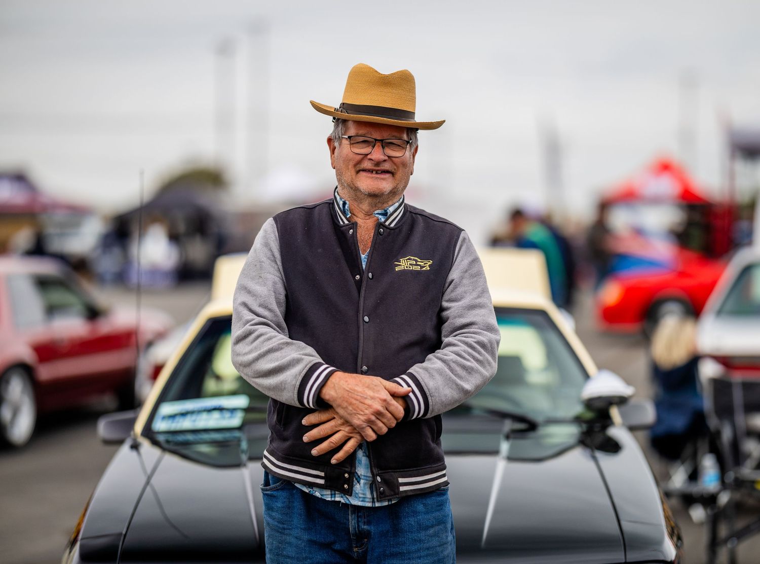 A person in a varsity jacket and brown hat stands in front of classic cars at an automotive event.