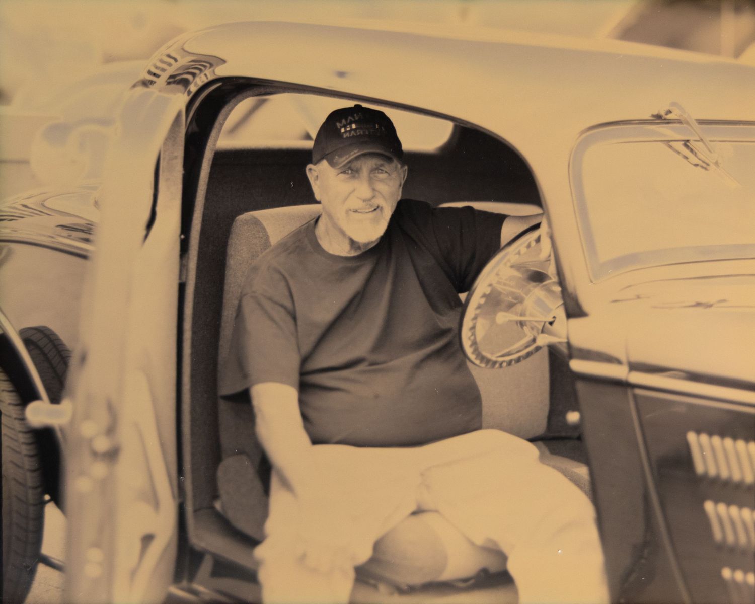 Older man in a baseball cap and t-shirt sitting in the driver seat of a vintage classic car.