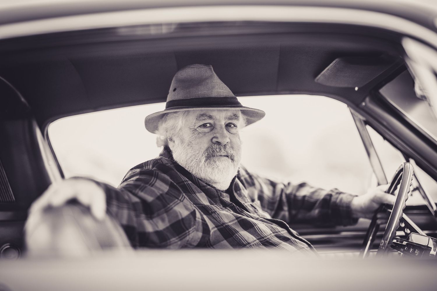 Elderly man with beard and hat driving a vintage car, black and white portrait photography.