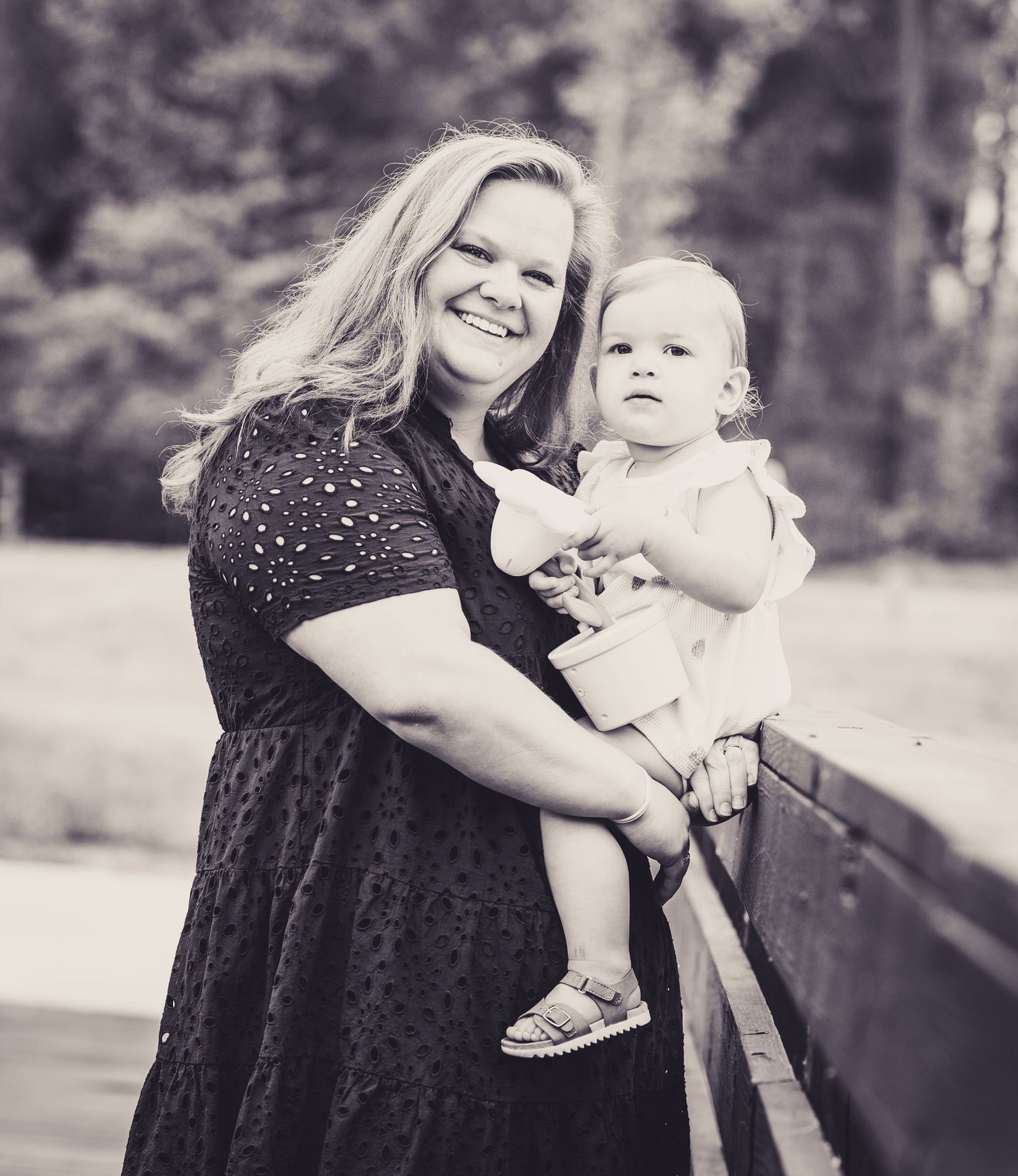 Black and white photo of a smiling woman holding a baby in white outfit, standing on a wooden bridge outdoors.