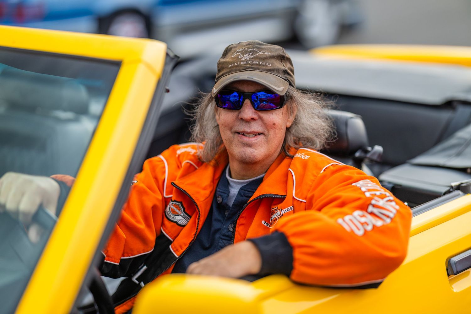 Person in orange racing jacket and blue sunglasses smiling in yellow convertible sports car at racing event.