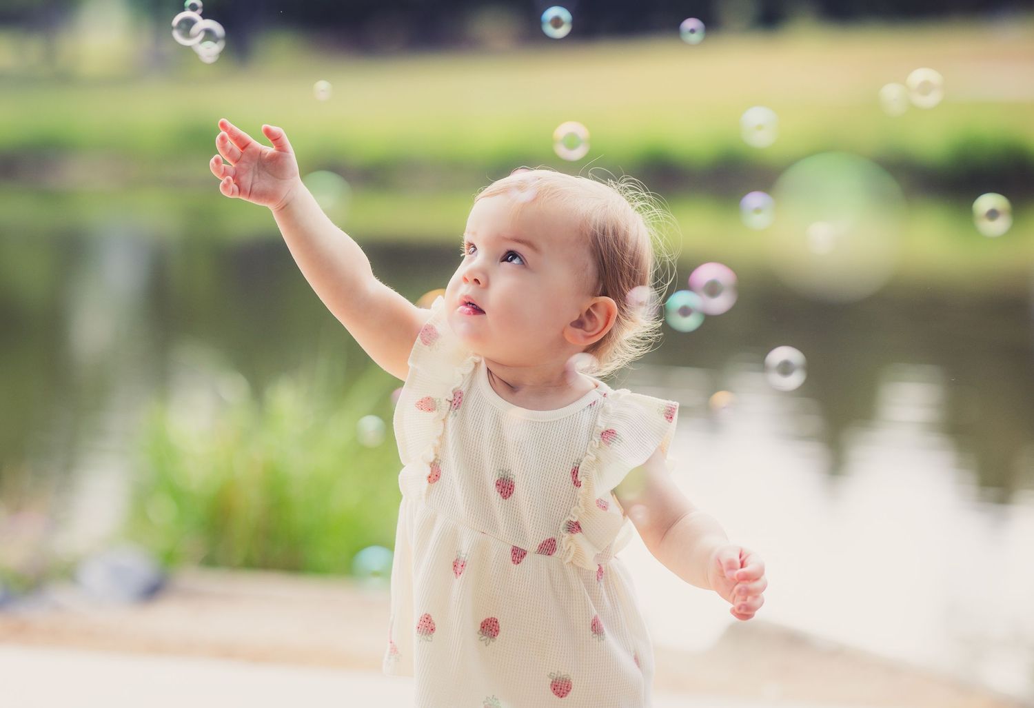 Adorable baby girl in white dress reaching for soap bubbles outdoors near a pond on a sunny day.