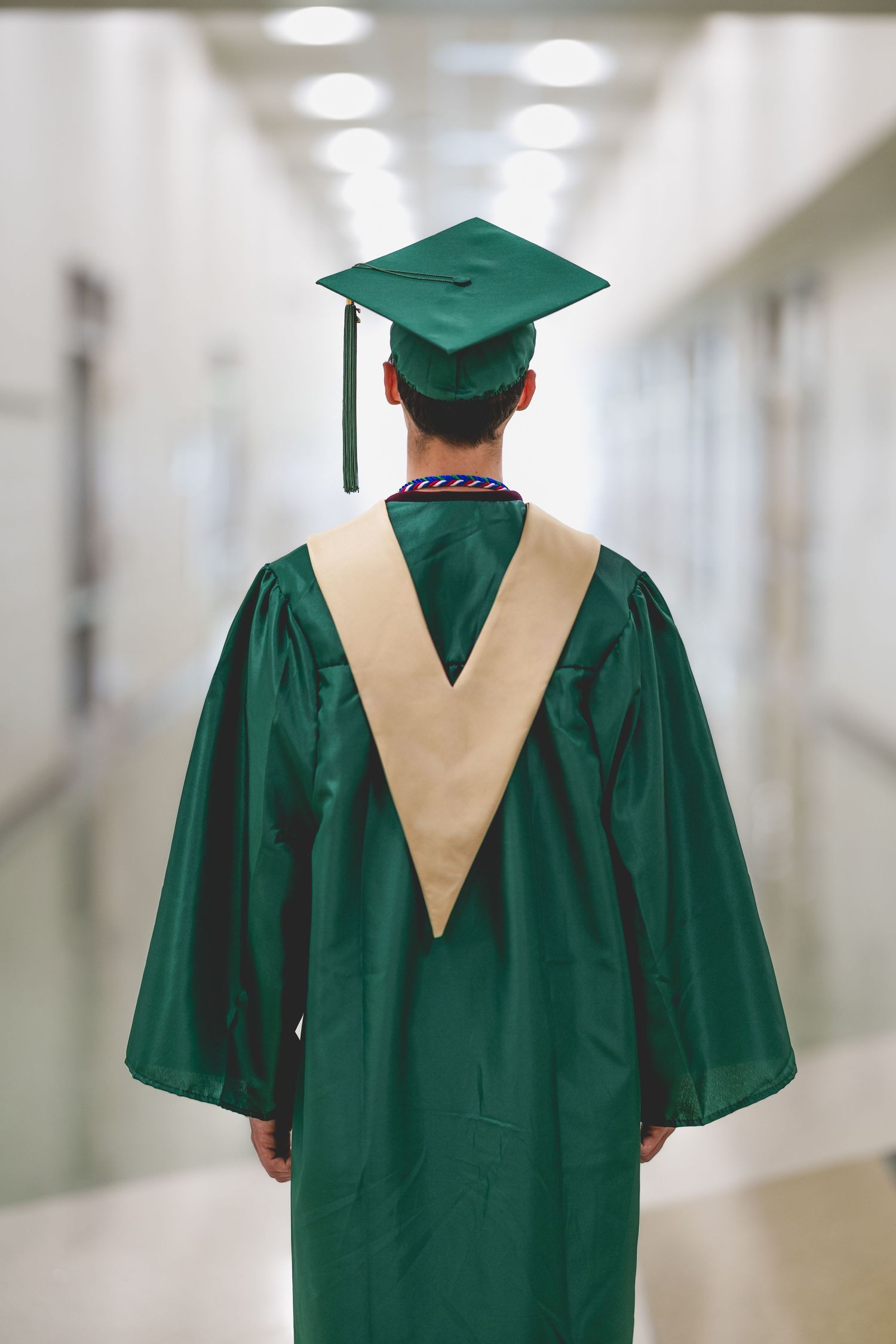 Graduate in green cap and gown stands in hallway, viewed from behind, wearing honor cords.