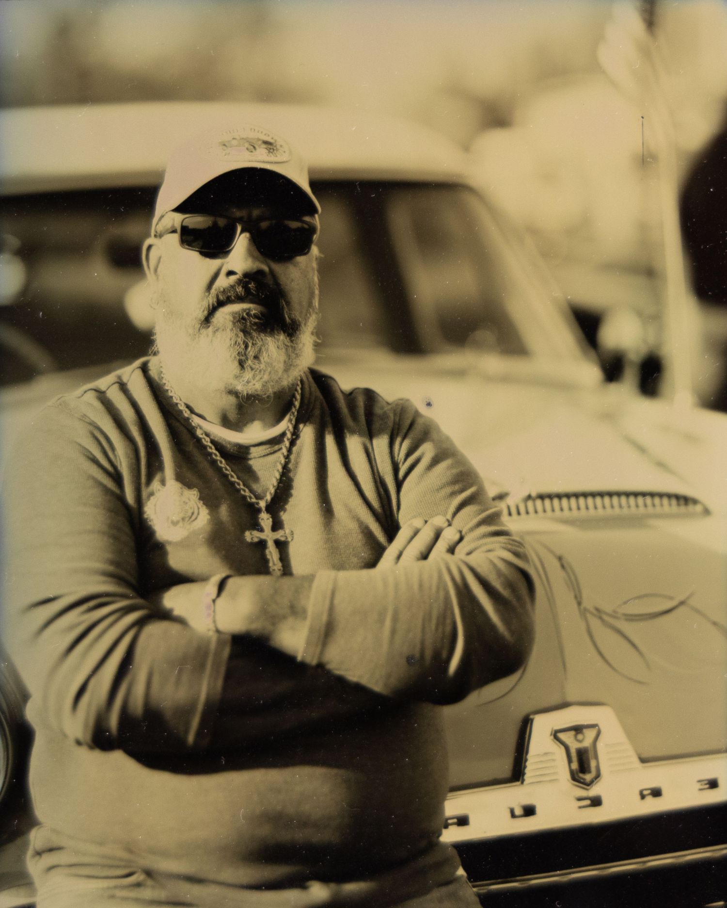 Older man with sunglasses, cap, and cross necklace leaning on vintage car in sepia-toned photo.