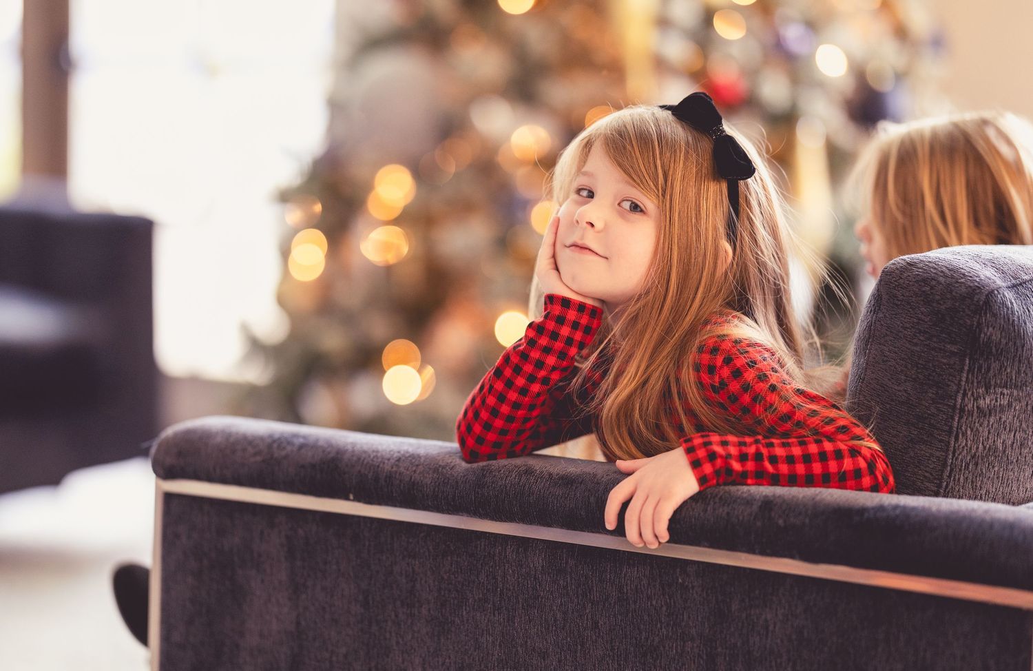 Young girl in red plaid outfit resting on sofa near decorated Christmas tree.