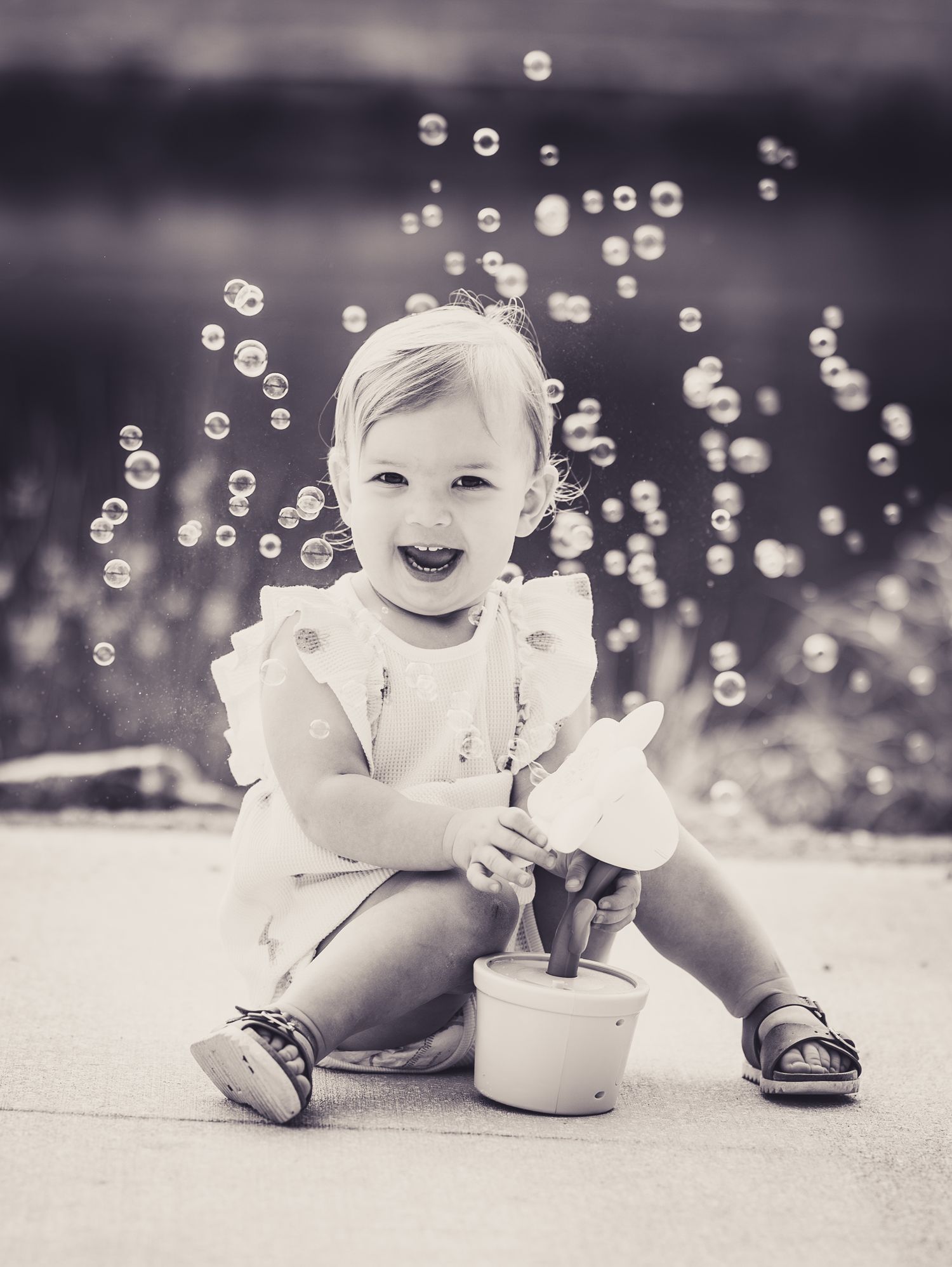 Black and white photo of a happy toddler girl sitting outdoors playing with bubbles in a white dress.