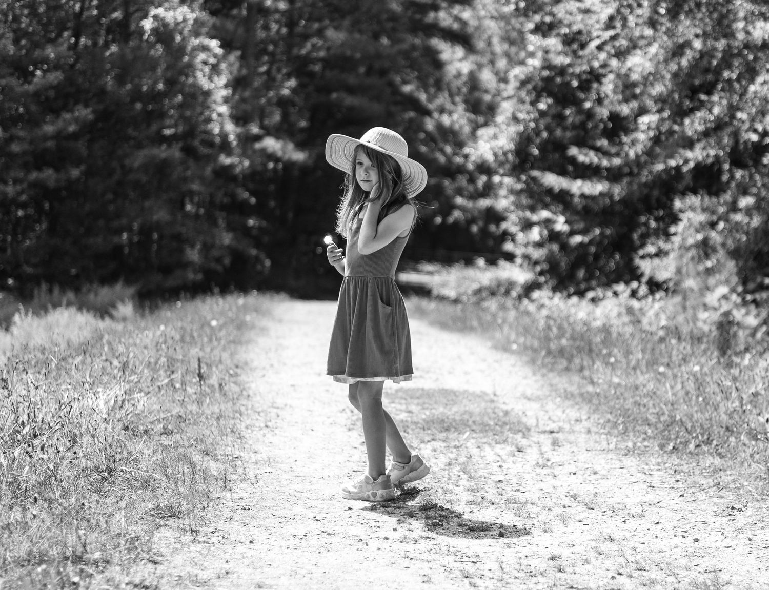 Black and white photo of a young girl in a dress and wide-brim hat standing on a dirt path surrounded by trees.