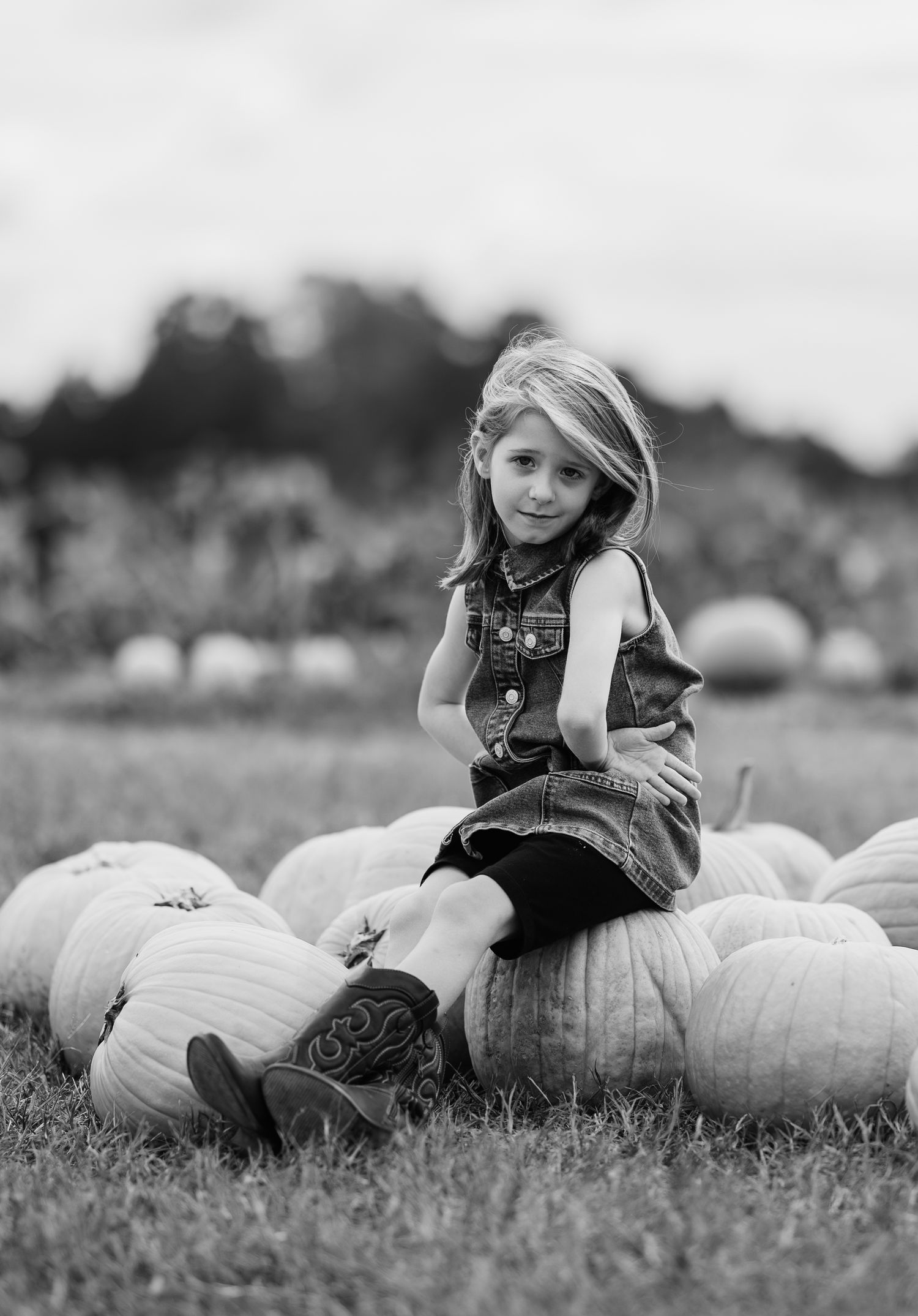 Young girl sitting on pumpkins in a field, wearing cowboy boots and denim vest, black and white photo.