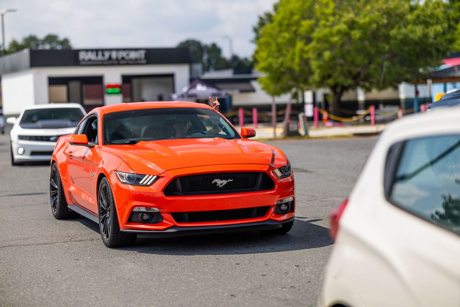 Bright orange Ford Mustang GT parked in a lot near a Rally Point store on a sunny day.
