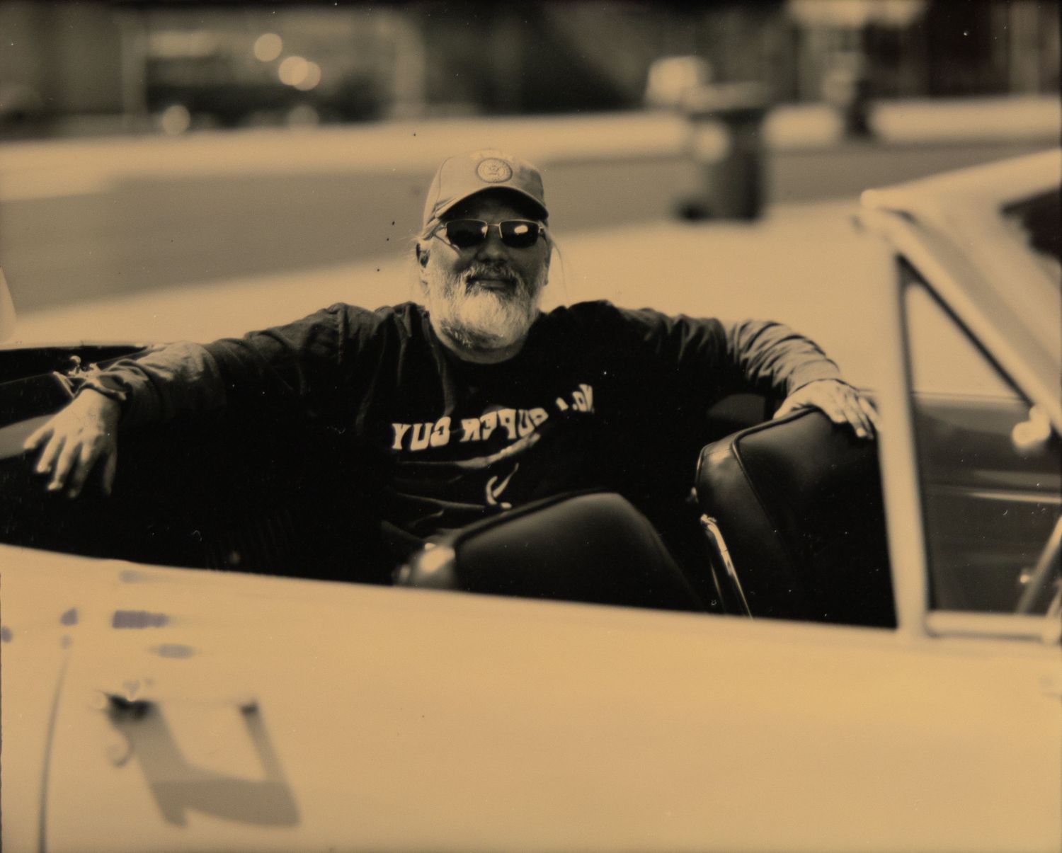 Older bearded man in cap and dark shirt relaxing in a classic white convertible car, sepia-toned photo.