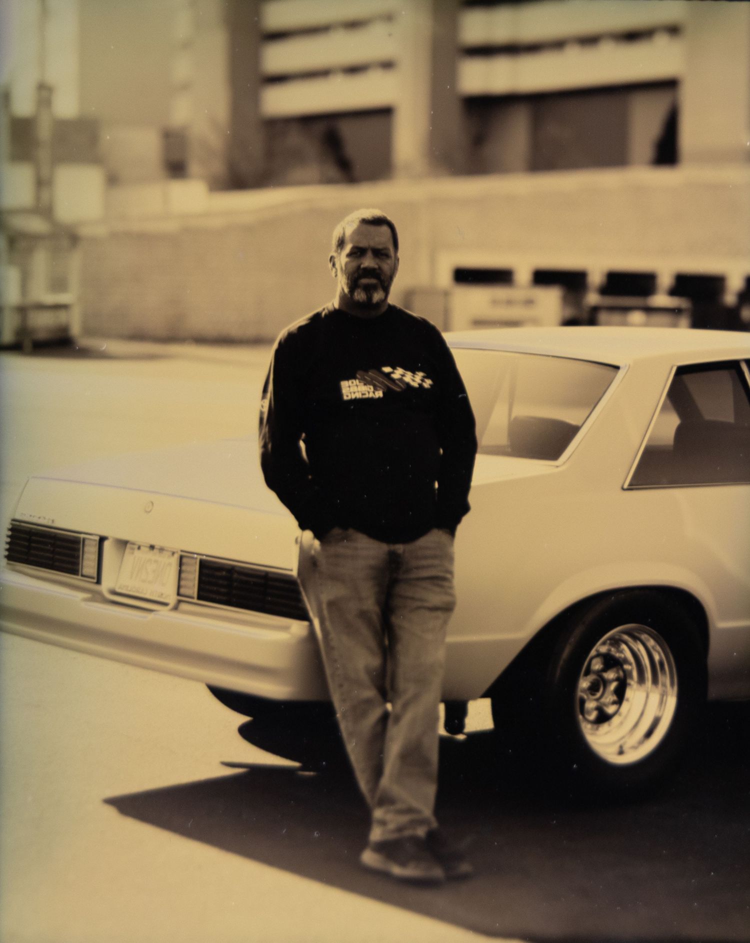 Man in black hoodie leaning against a classic white muscle car in an urban parking lot, sepia-toned photo.