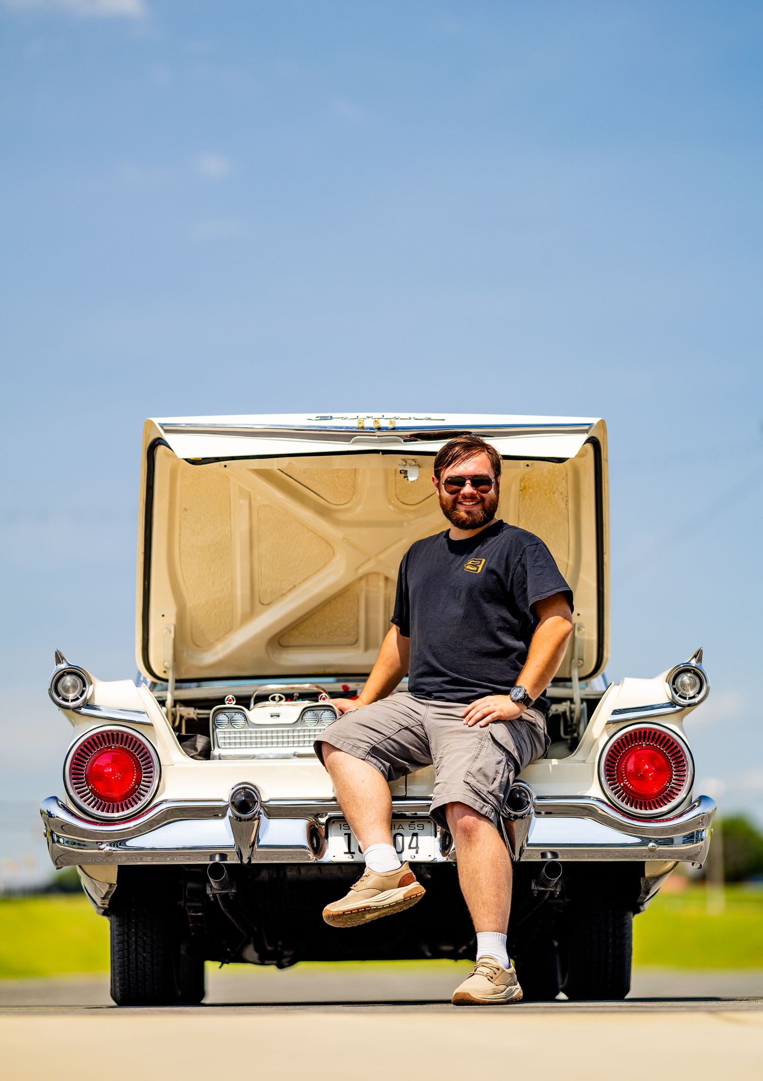 Man sitting on rear bumper of classic white vintage car with open trunk on sunny day outdoors.