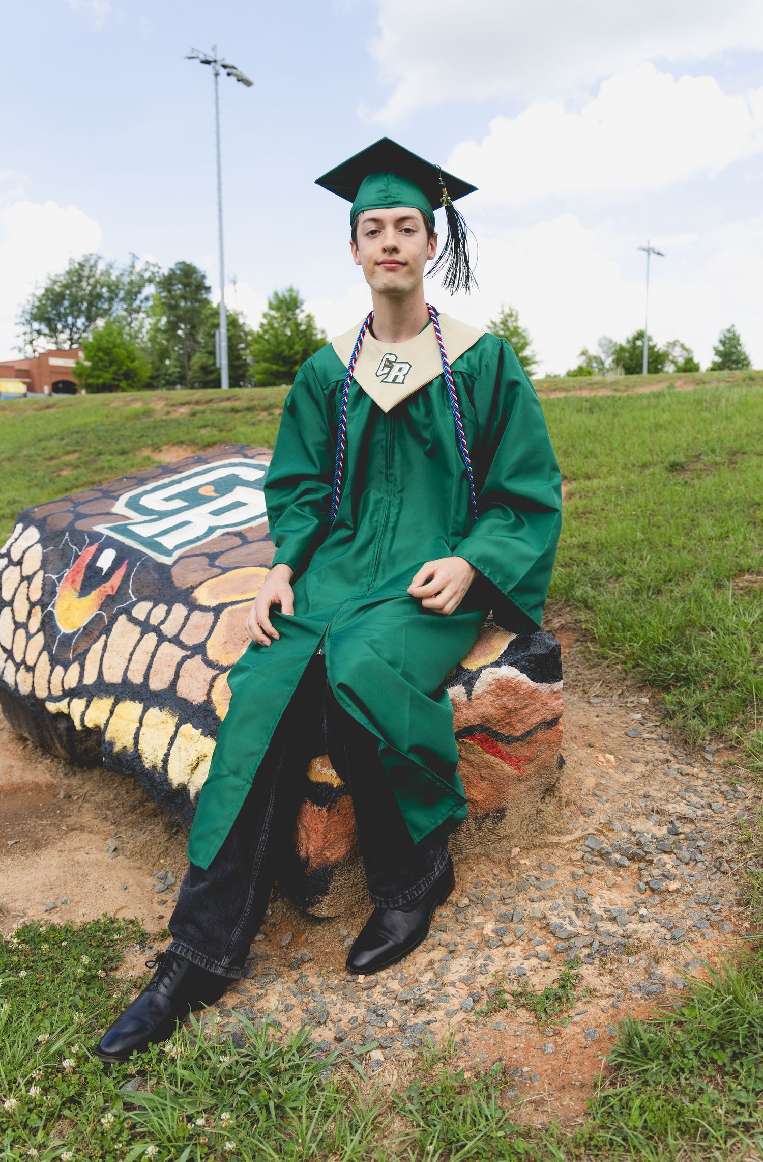 Graduate in green cap and gown sits on painted snake sculpture outdoors, wearing black boots and honor cords.