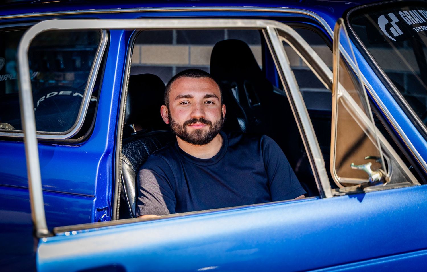A smiling man sits in the driver's seat of a classic blue car with a roll cage visible inside.