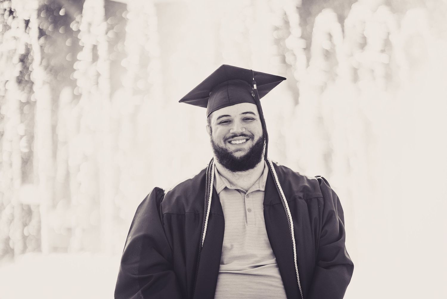 Smiling graduate in cap and gown poses outdoors in black and white photo near a fountain.