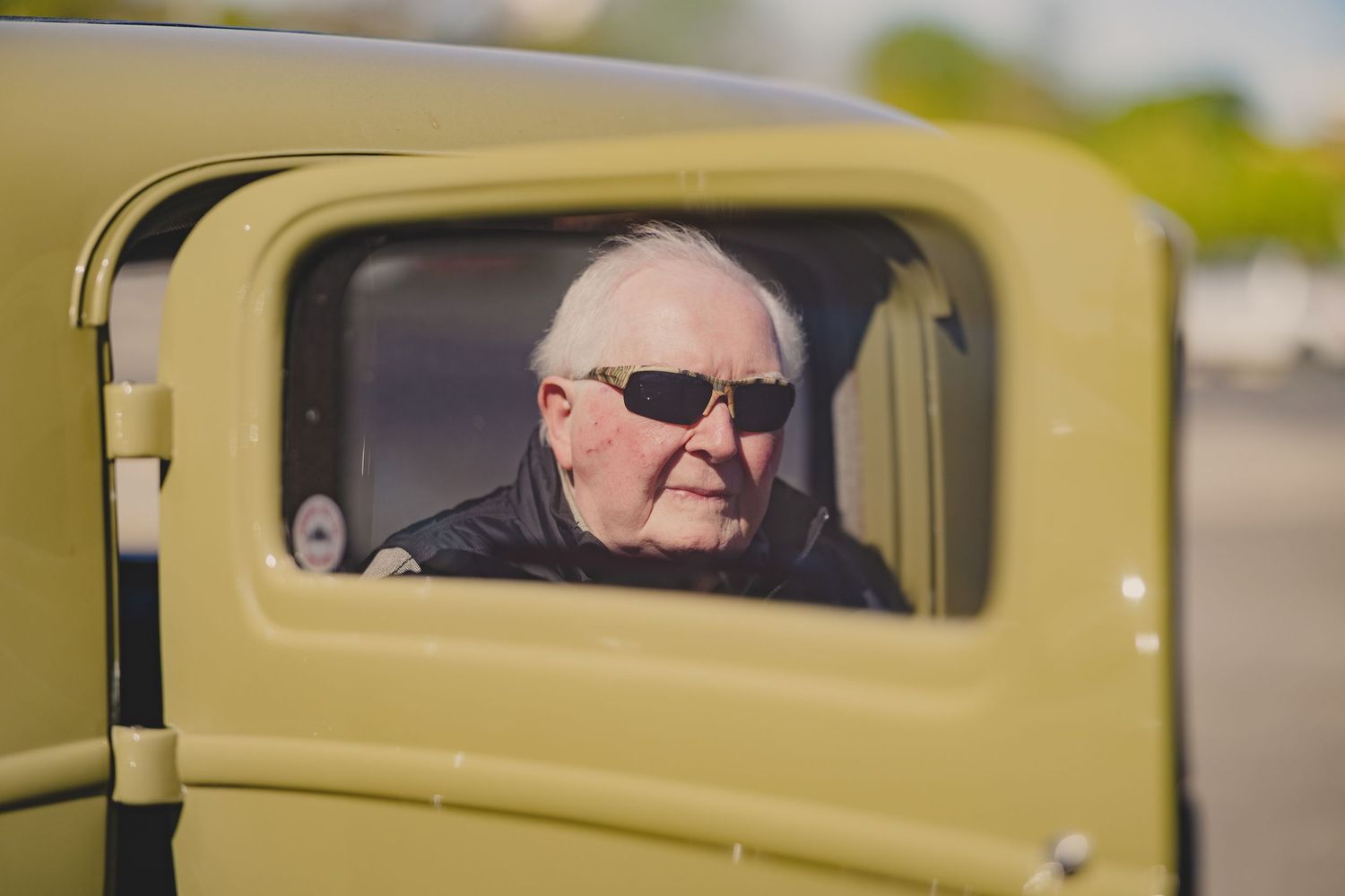 Elderly man in sunglasses seen through the window of a vintage olive-green classic car.
