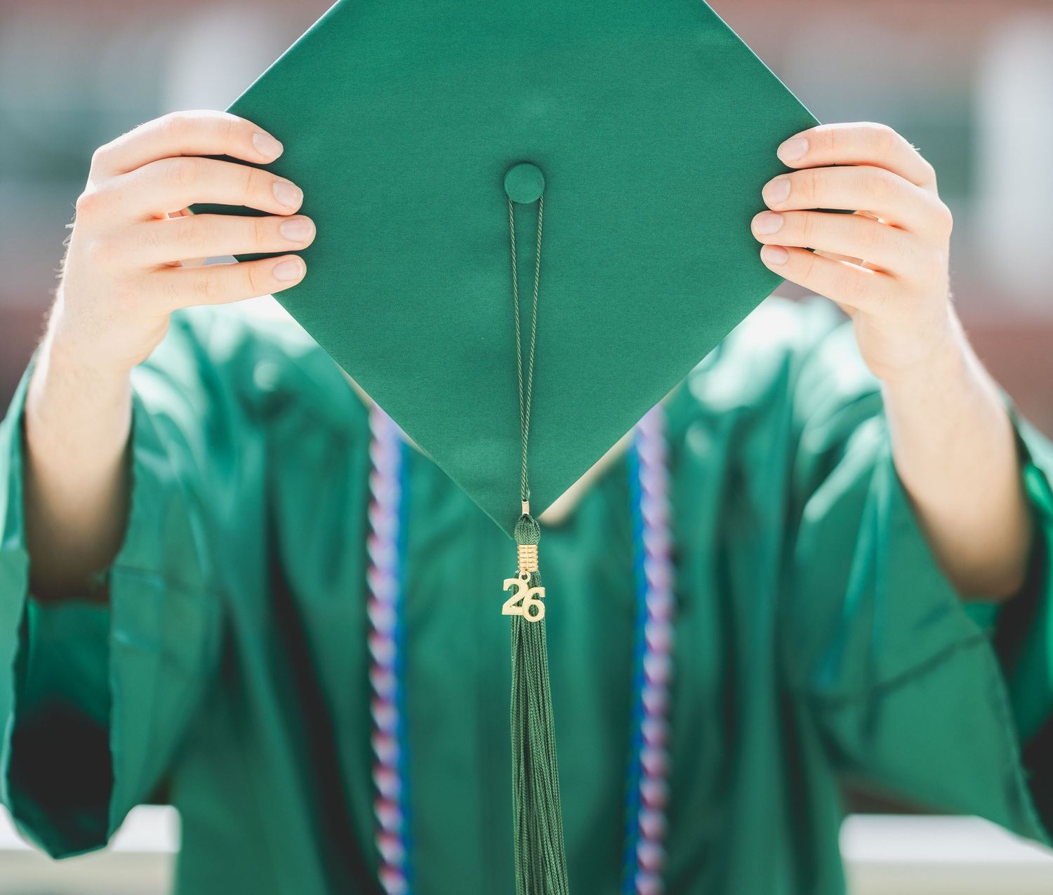 Student in green graduation cap and gown holding mortarboard up with gold '26 tassel for class of 2026.