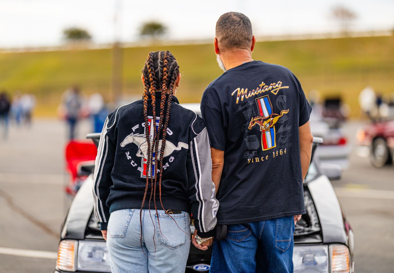 Two people wearing matching black t-shirts stand together looking at a car at an outdoor event.