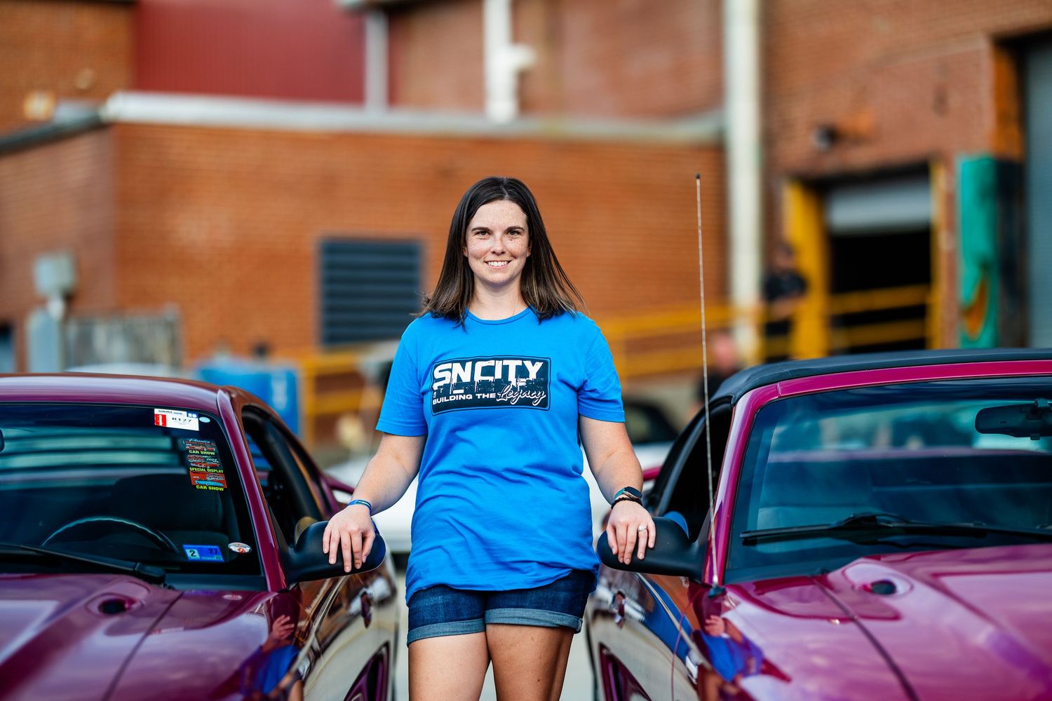 Woman in blue Snotty shirt stands between two classic cars with open hoods in a parking lot.