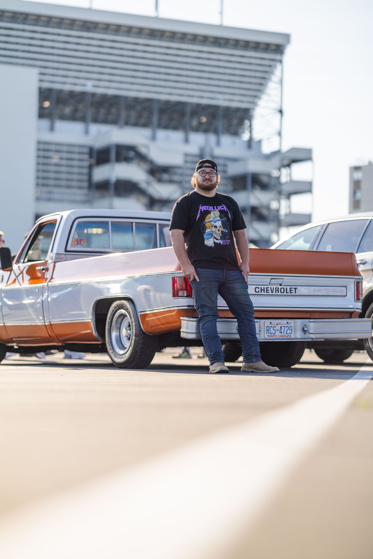 Man in Metallica shirt stands proudly beside a classic Chevrolet truck at an outdoor car show.