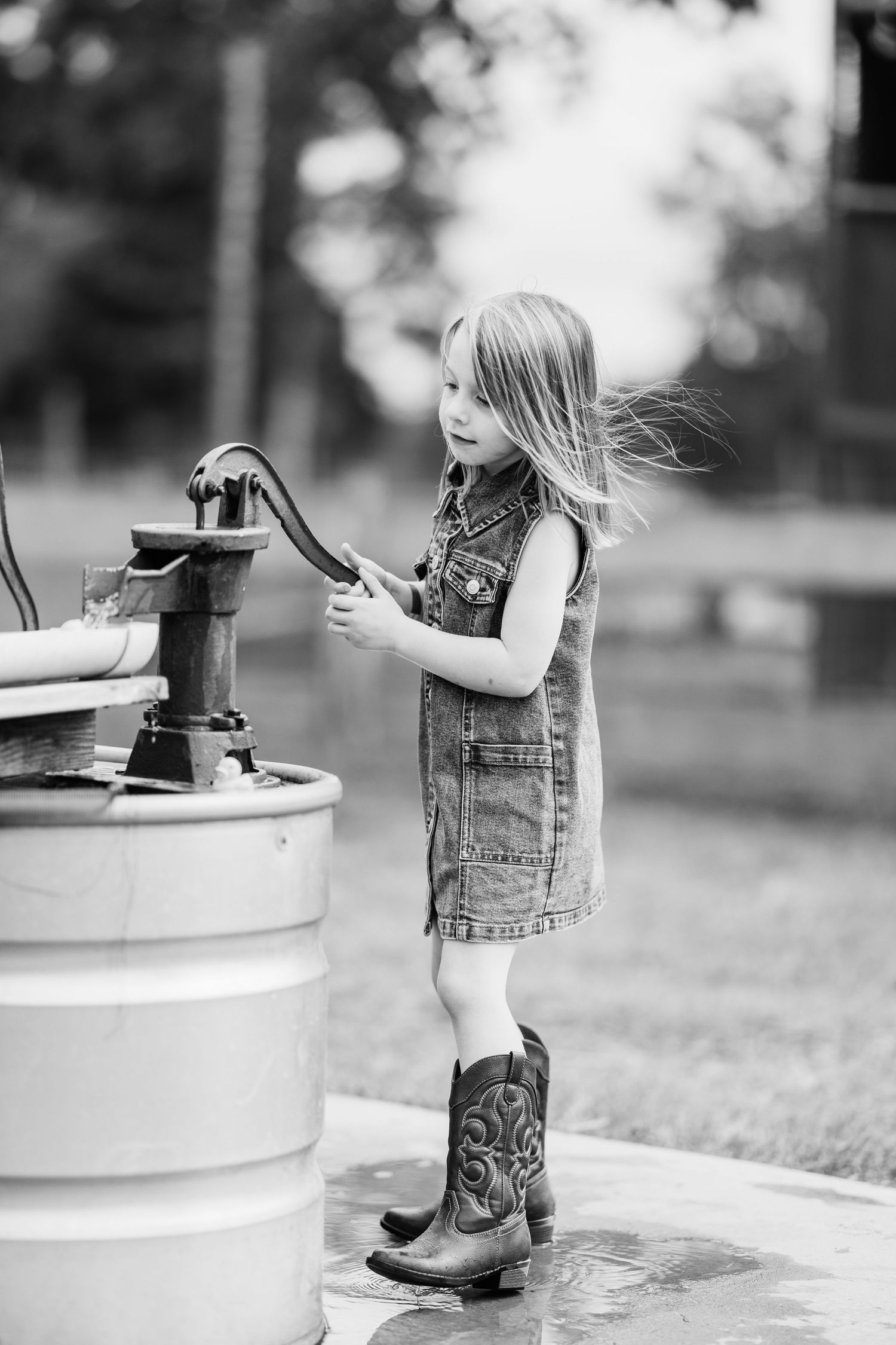 Black and white photo of a young girl in denim dress and cowboy boots pumping water from an old hand pump outdoors.