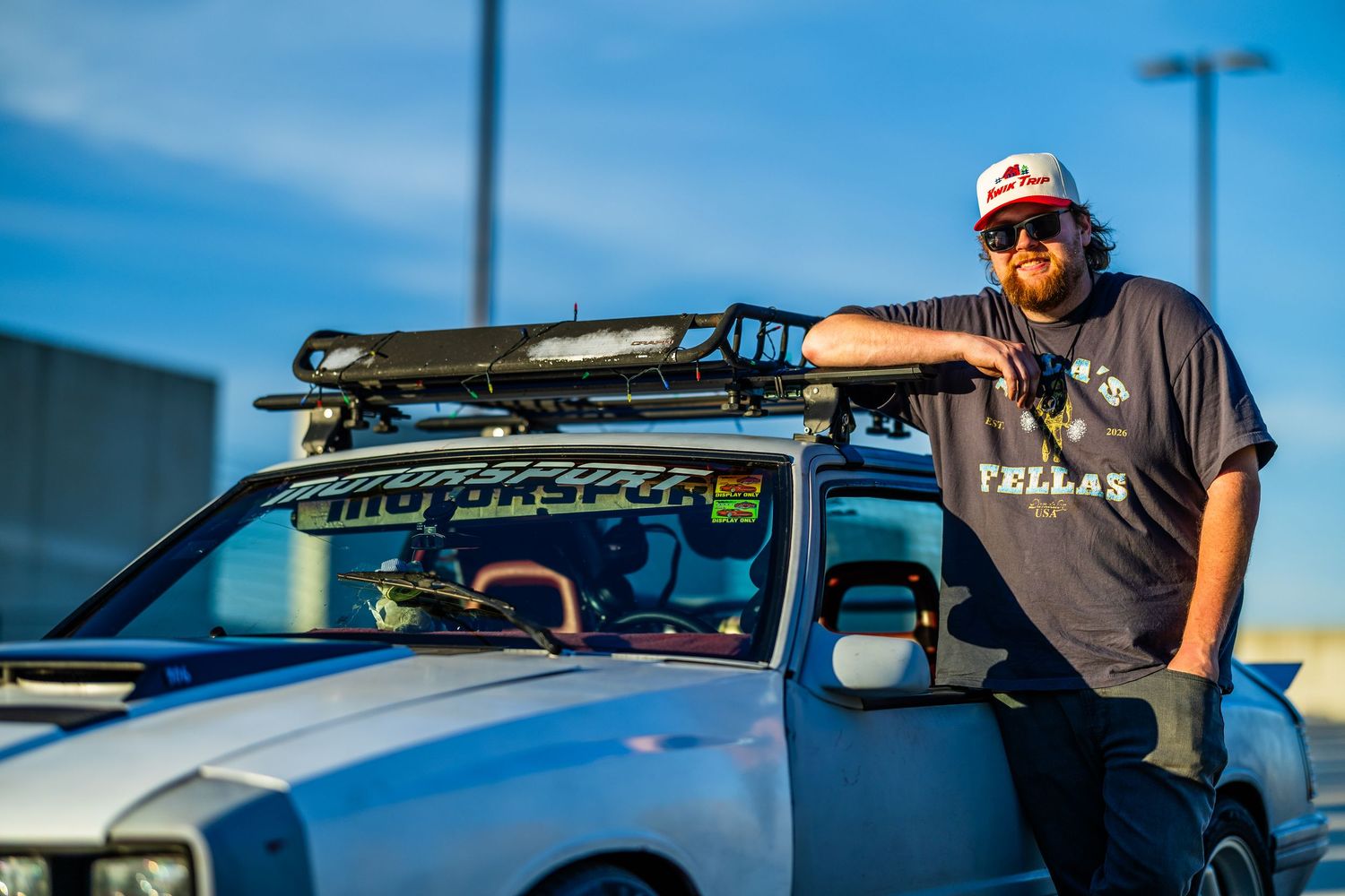 Man in trucker hat leaning on modified off-road truck with roof rack at dusk.