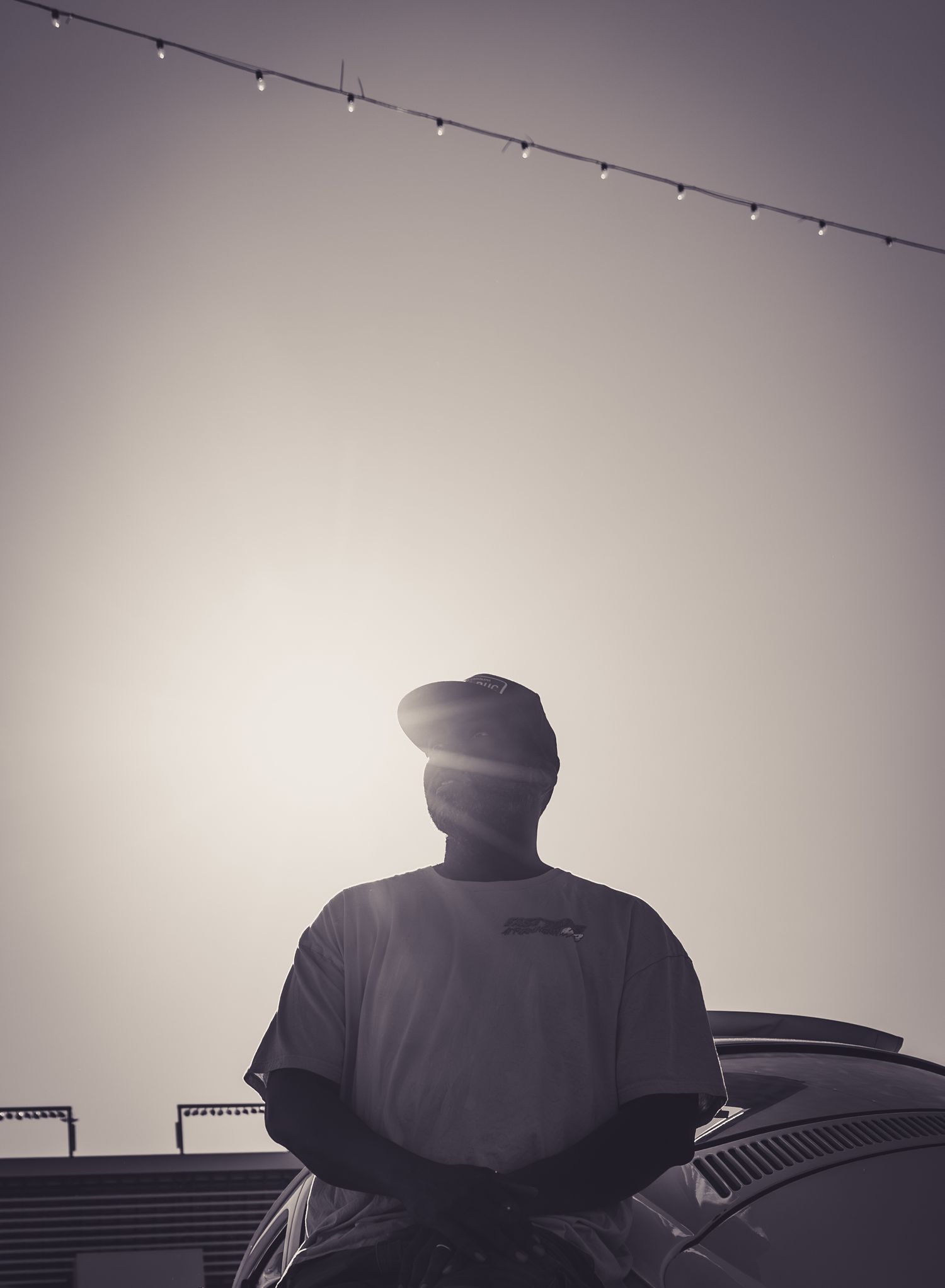 Silhouetted man wearing cap and t-shirt backlit by sun, string lights above, monochrome moody outdoor portrait.