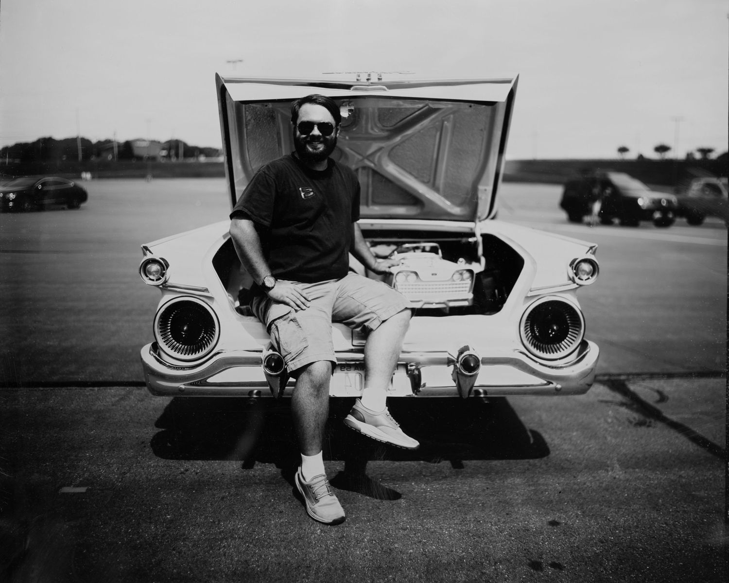 A bearded man in sunglasses sits on the trunk of a vintage white car with hood open, black and white photo.