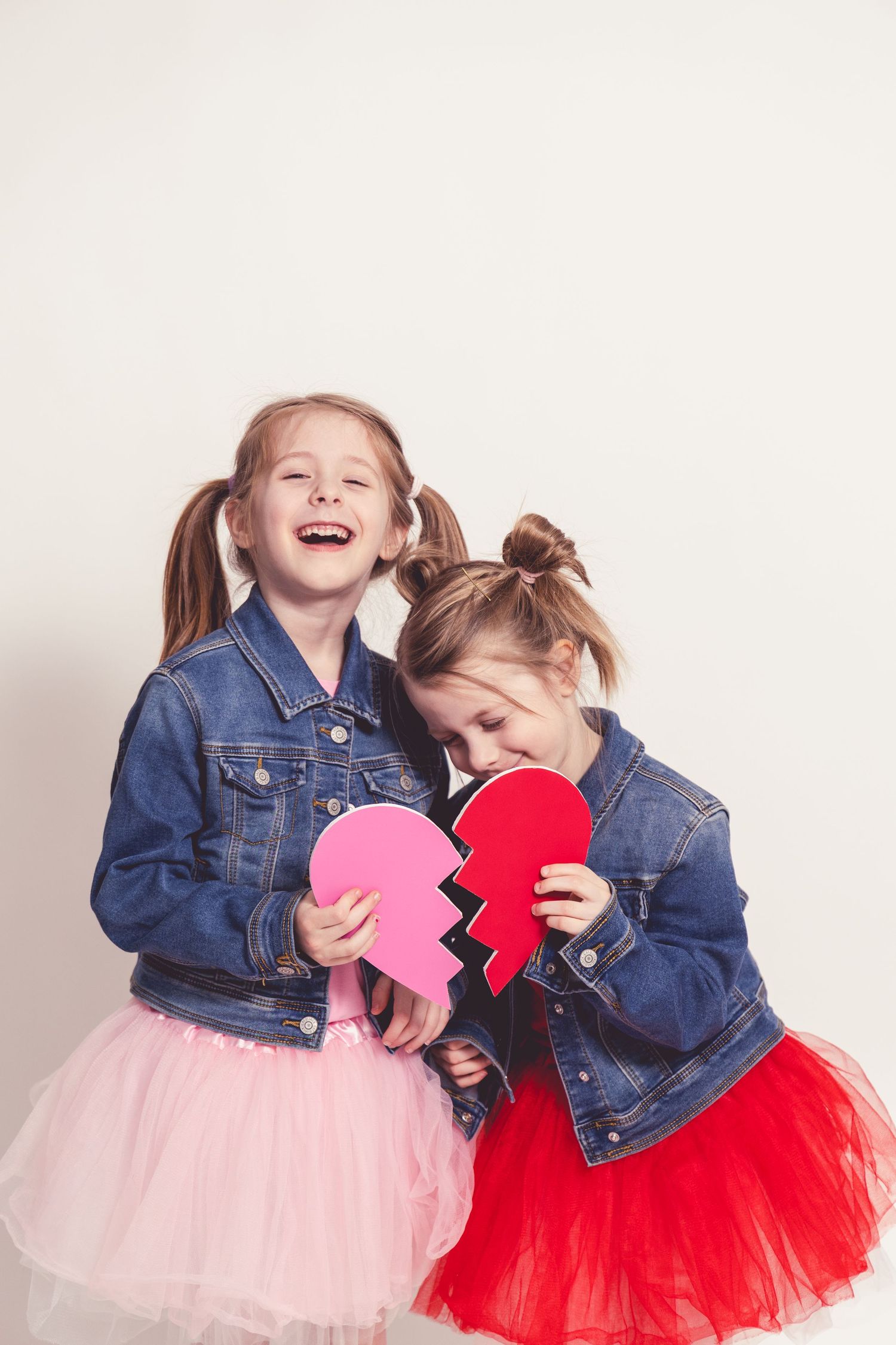 Two little girls in denim jackets hold a split paper heart, laughing together.