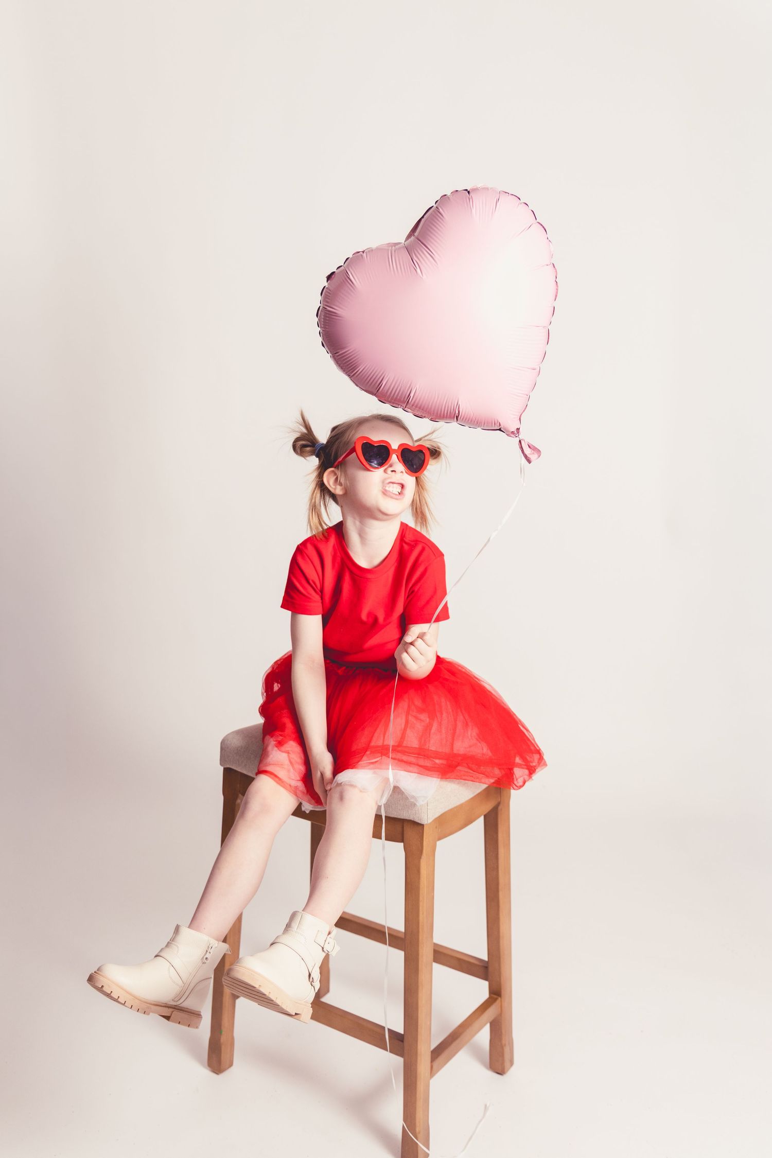 A person in a red dress and heart sunglasses sits on a wooden stool with a pink heart balloon against a white background.
