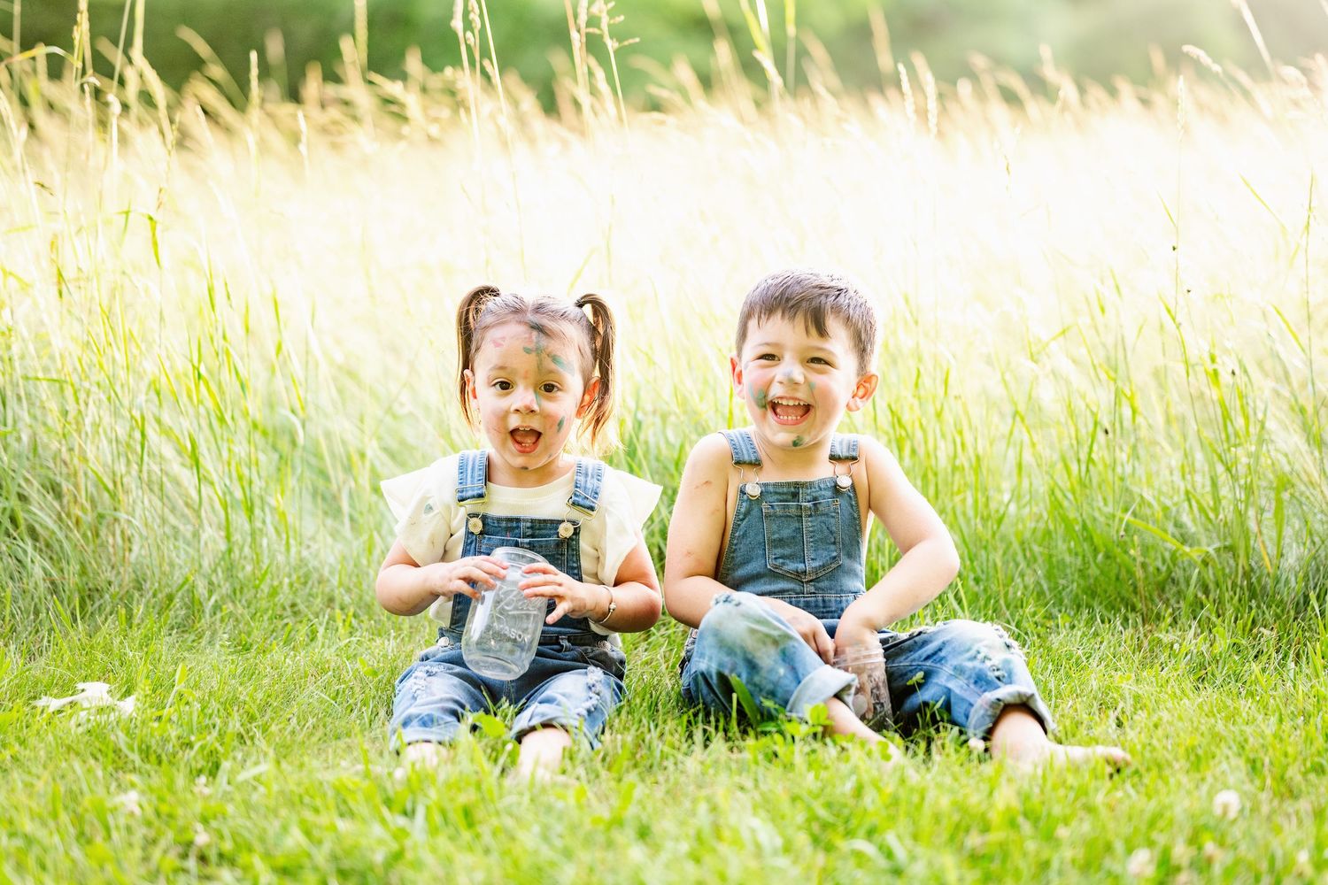 Two children in denim overalls sit together laughing in a sunlit meadow with tall grass.