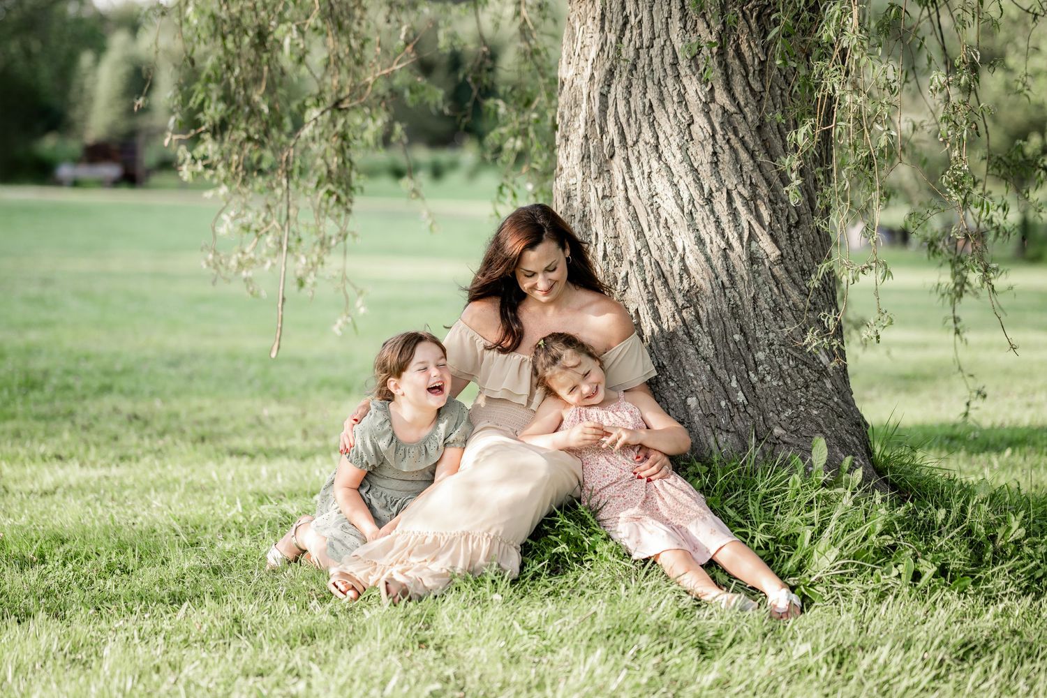 A couple sits together under a large tree in a peaceful grassy meadow on a sunny day.