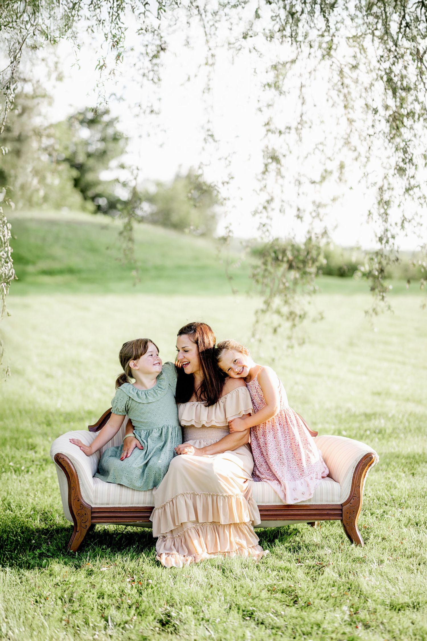 Three people share a tender moment while sitting on an antique chaise lounge beneath a weeping willow tree in a grassy field.