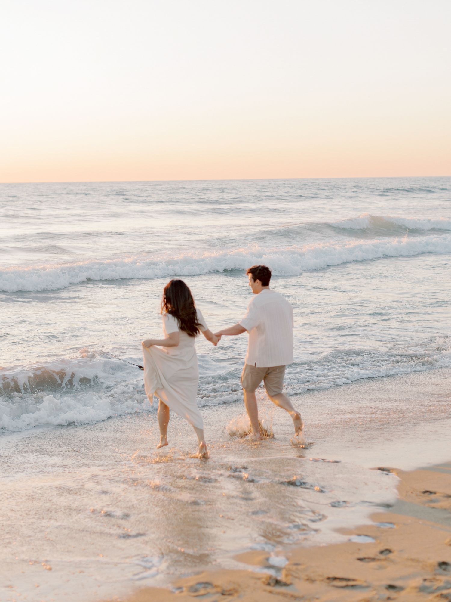 Couple holding hands and running on a sandy San Francisco beach at sunset with gentle waves in the background.