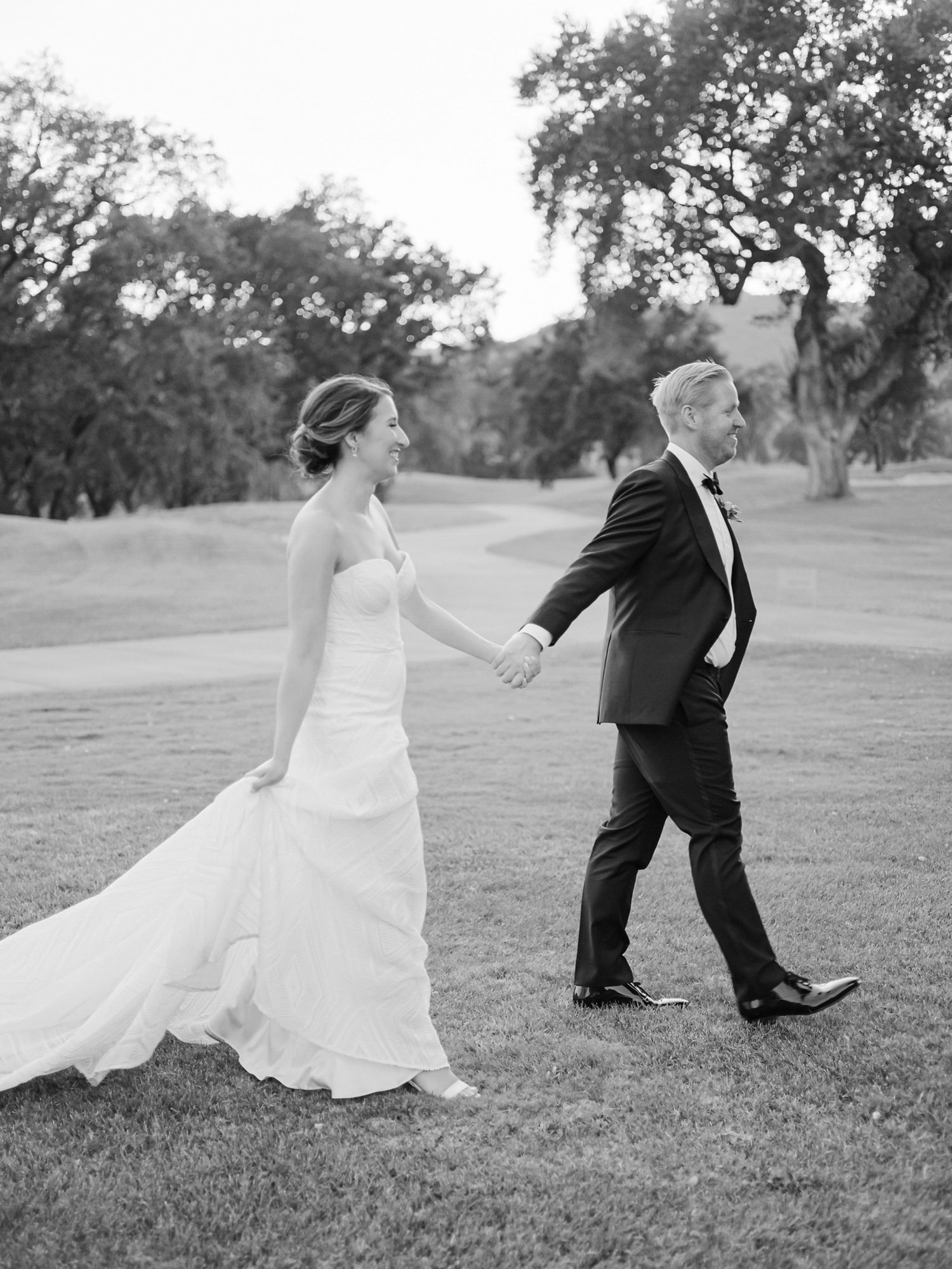 A couple in wedding attire walking together on a grassy field with oak trees in the background at Sonoma Golf Club.