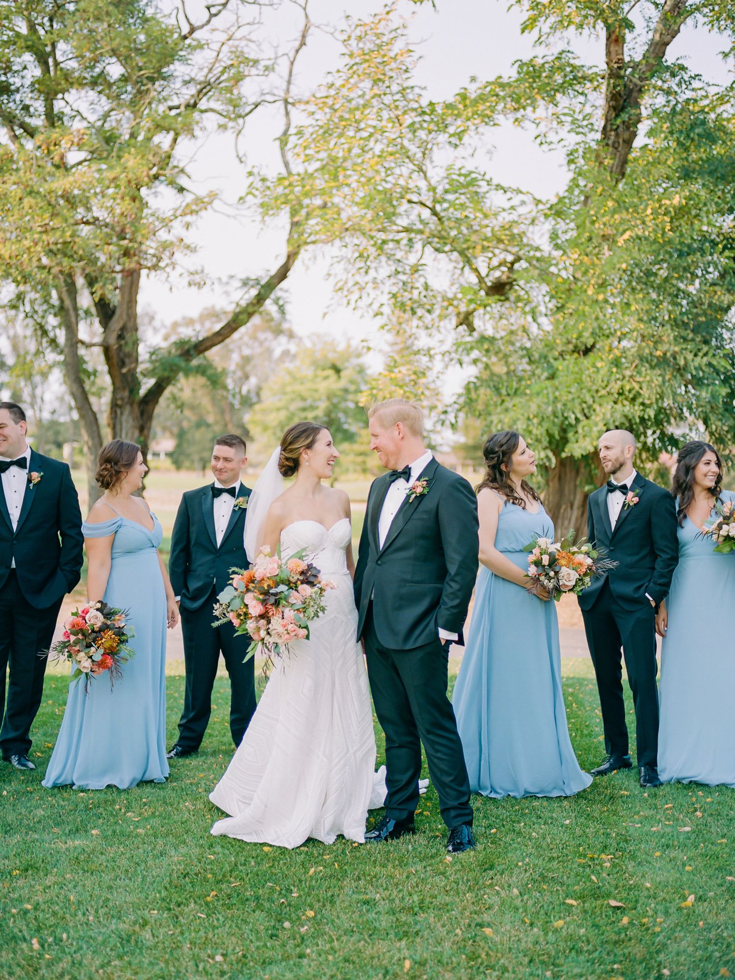 Wedding party in light blue bridesmaid dresses and black tuxedos poses outdoors under trees on a sunny day at Sonoma Golf Club.