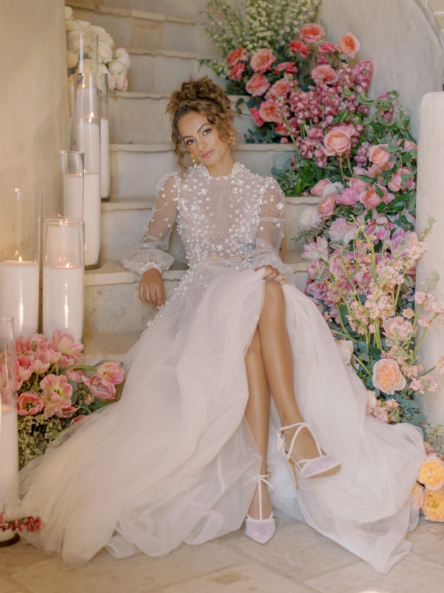 A bride in a romantic lace gown poses on marble stairs surrounded by pink floral arrangements at Sunstone Winery in Santa Ynez.