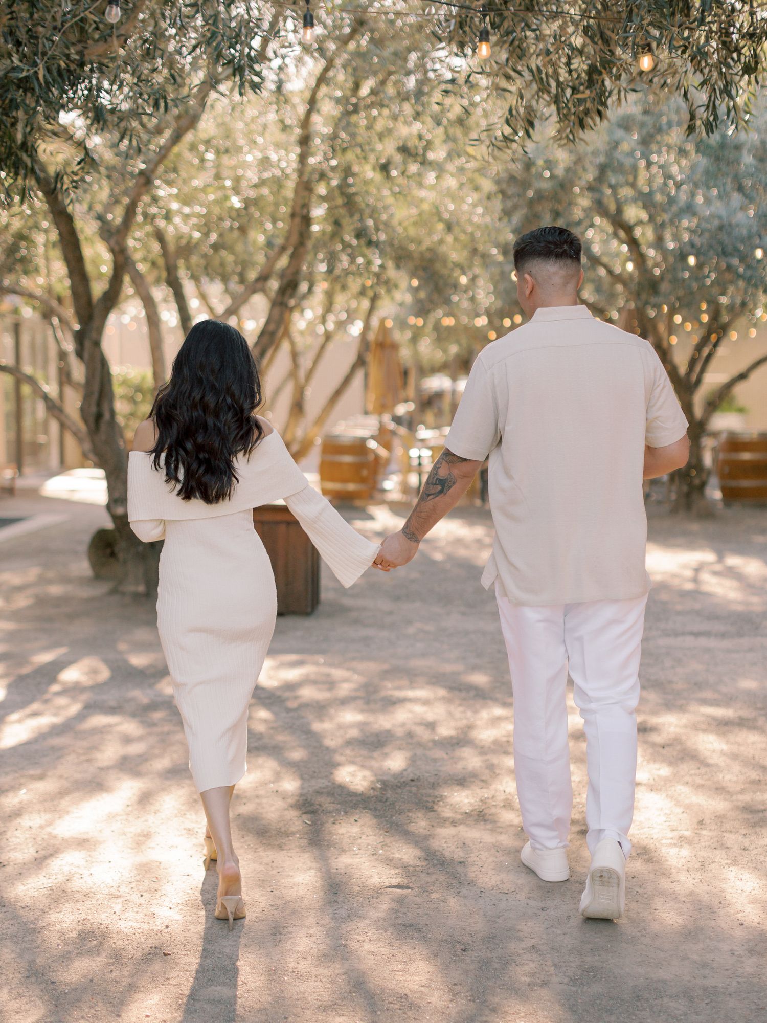 Couple in white outfits walking hand in hand through a sun-dappled tree-lined path at Cornerstone Sonoma.