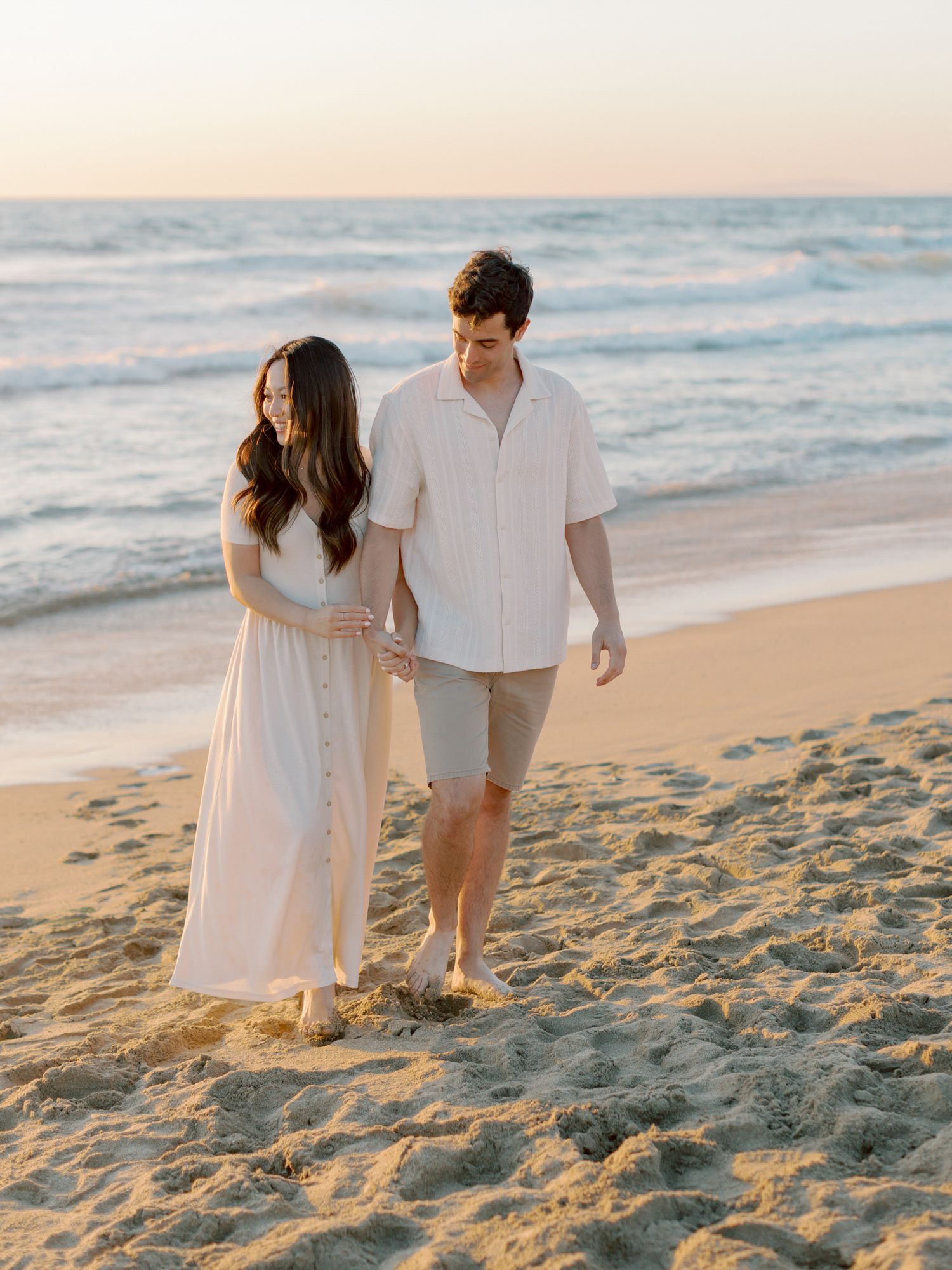 Couple walking together on San Francisco beach at sunset wearing white casual outfits.