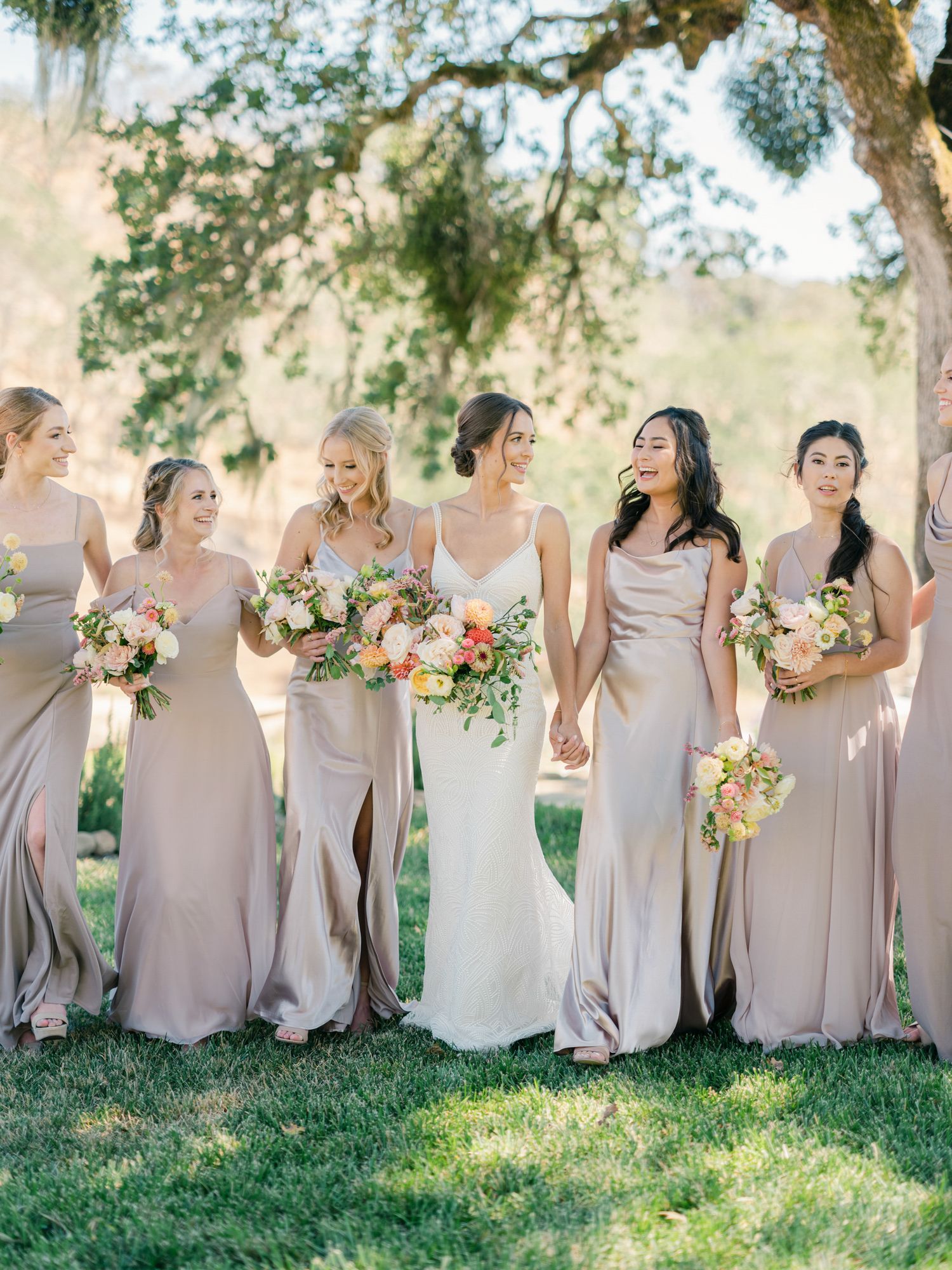 Bridesmaids in blush pink dresses holding colorful bouquets while standing on a sunny lawn in Napa.