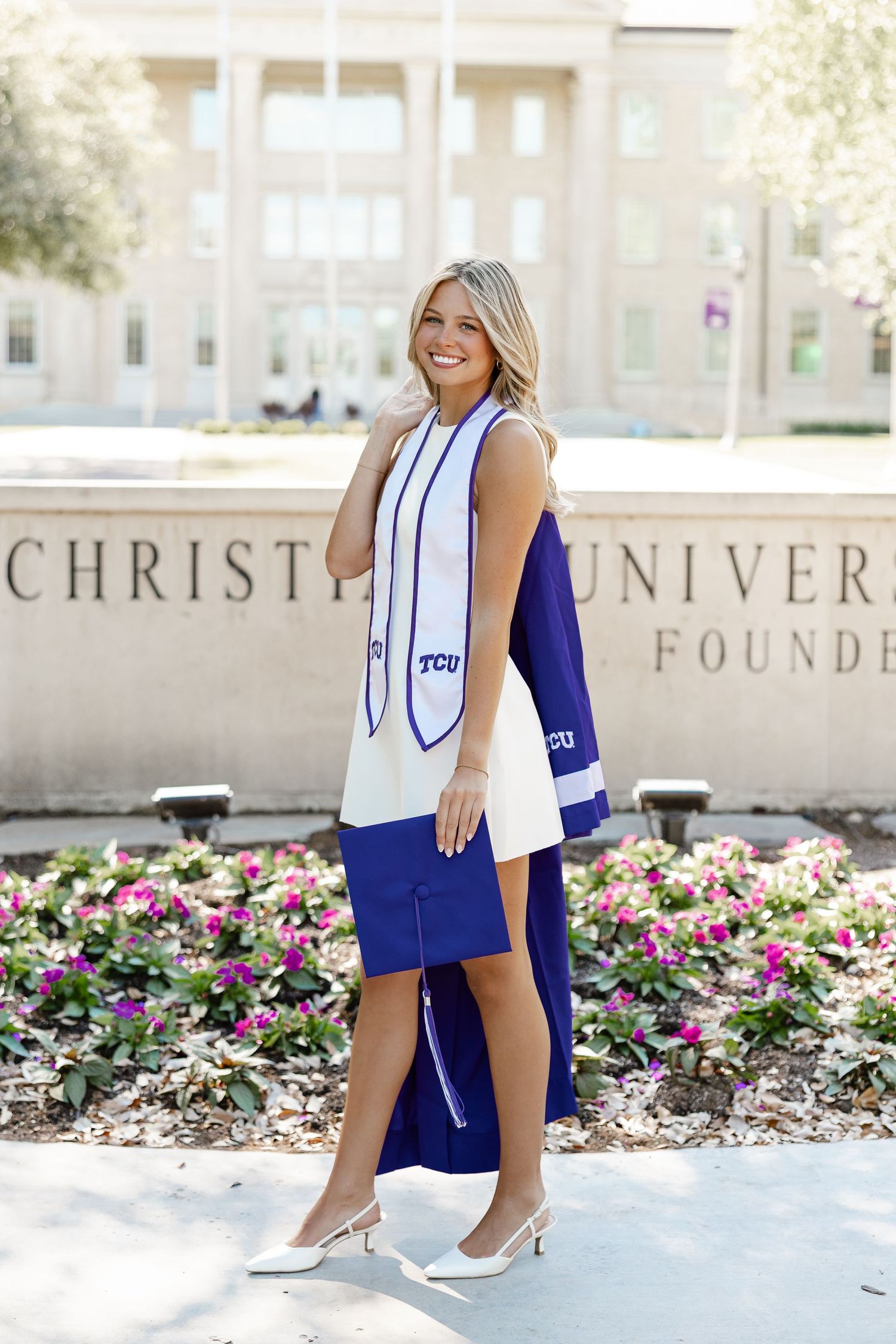 Smiling TCU graduate poses in white dress with purple regalia outside Texas Christian University.