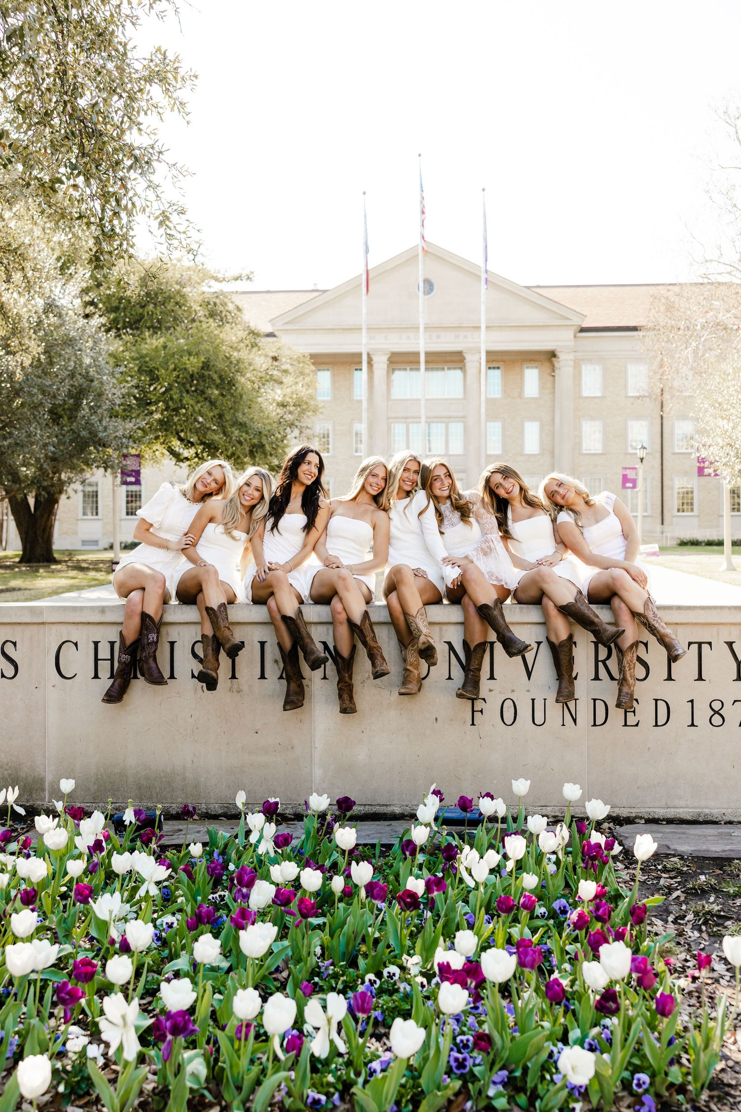 Group of women in white dresses and boots sitting on Texas Christian University founded sign with tulips.