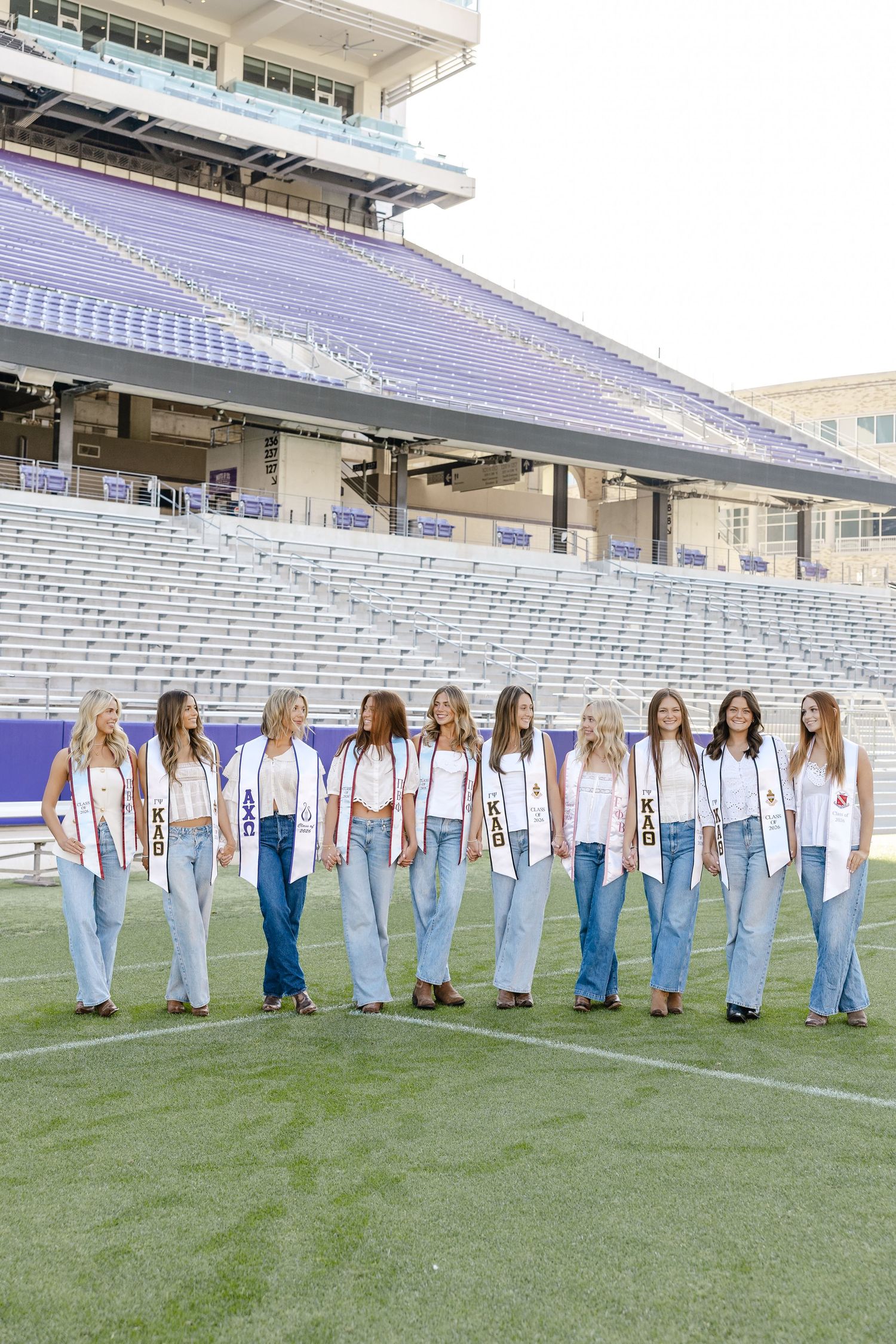 Group of sorority women in white vests and jeans posing on a college football field with purple stadium seats.