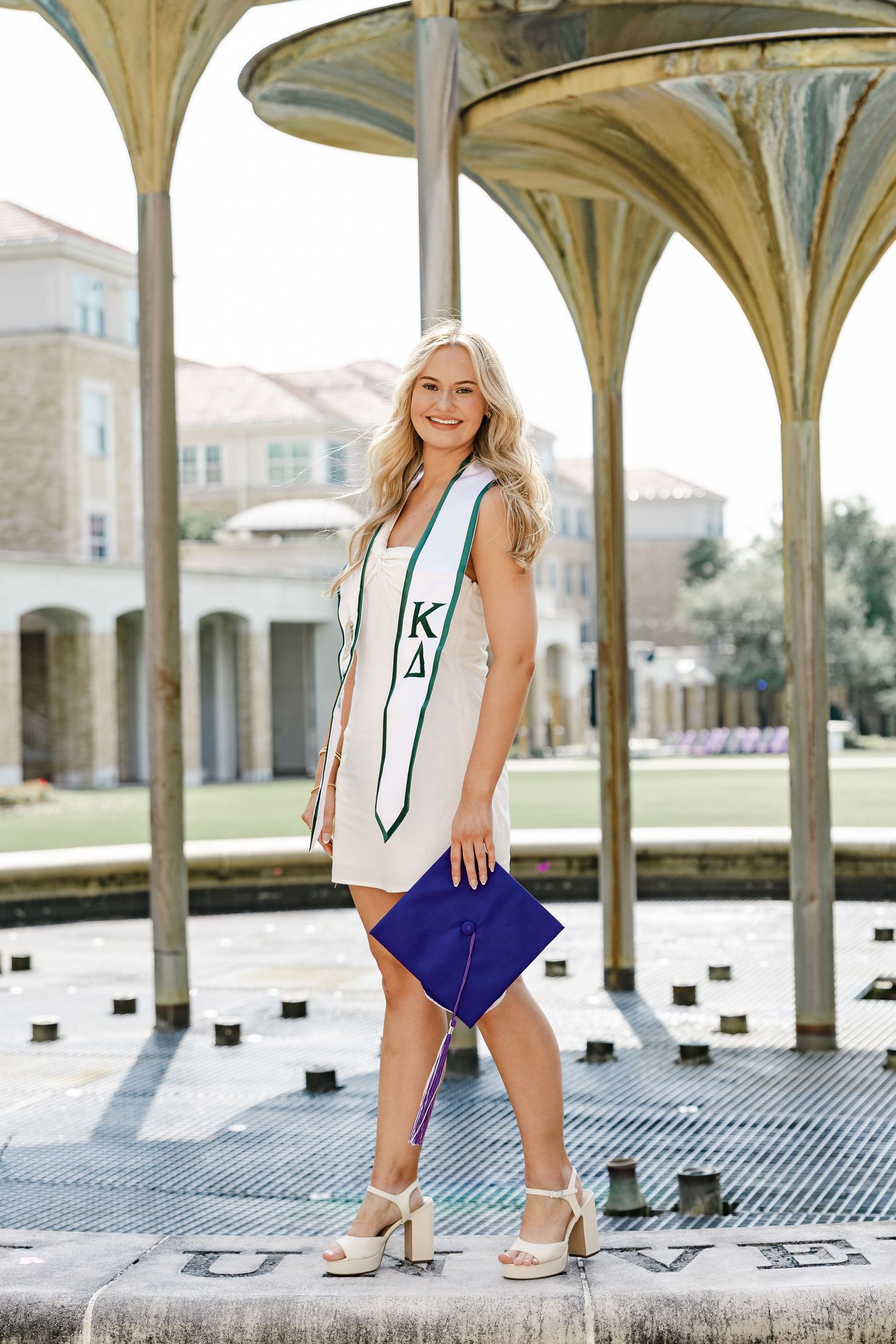 Smiling blonde graduate wearing Kappa Delta stole holds blue cap at outdoor campus pavilion.