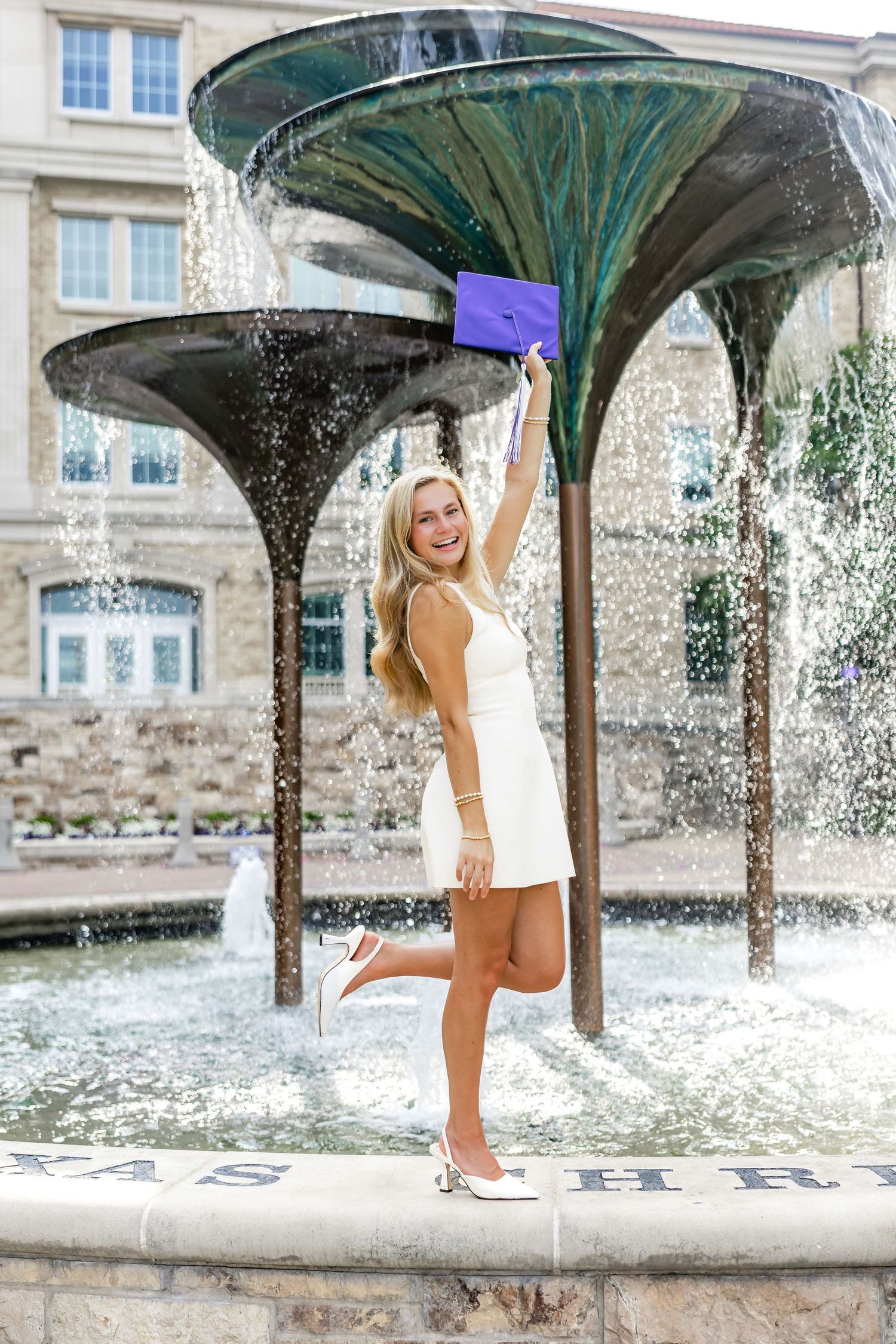 Graduate celebrates by fountain holding diploma in white dress at university.