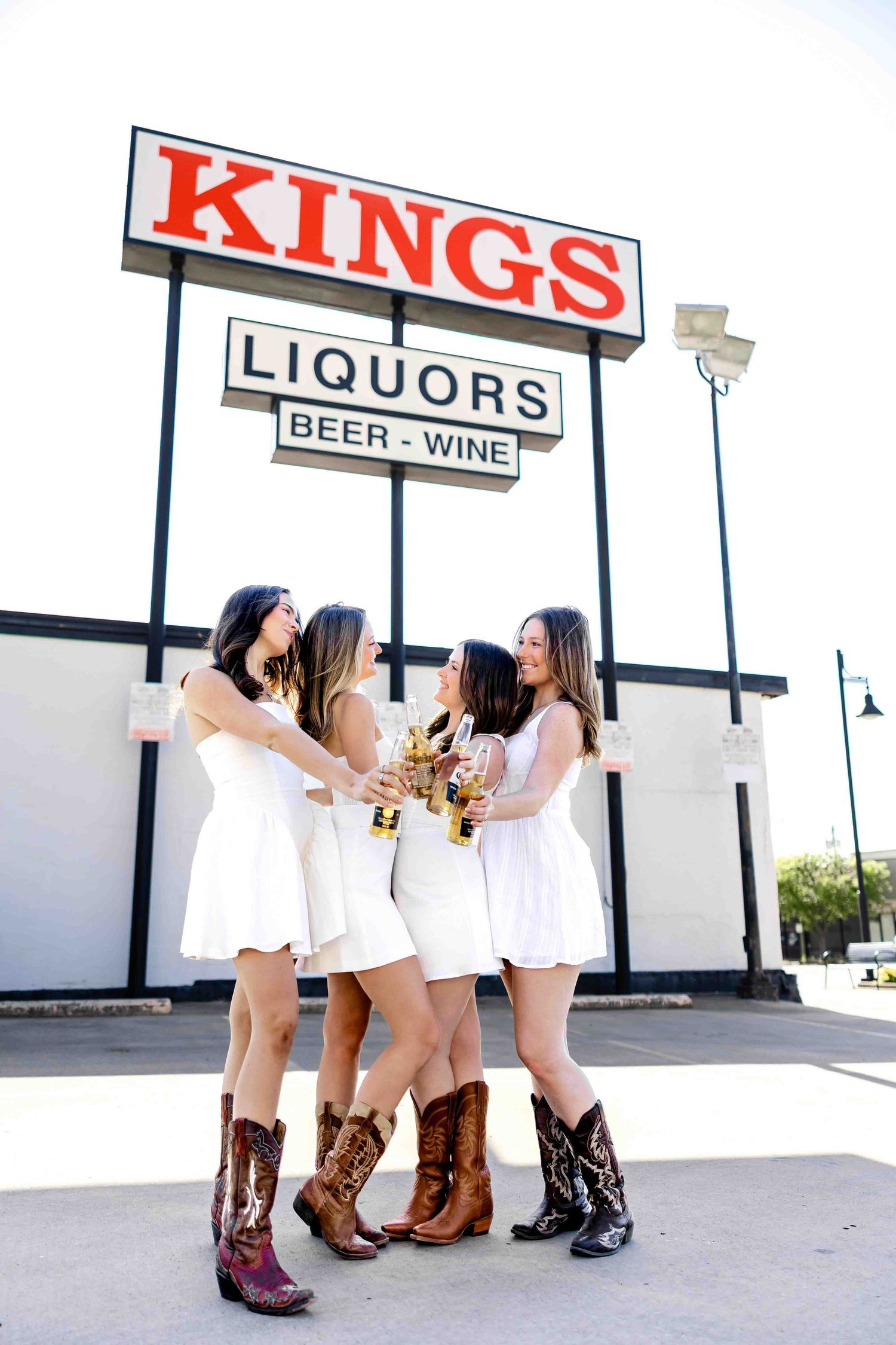 Group wearing white dresses and cowboy boots poses outside a liquor store.