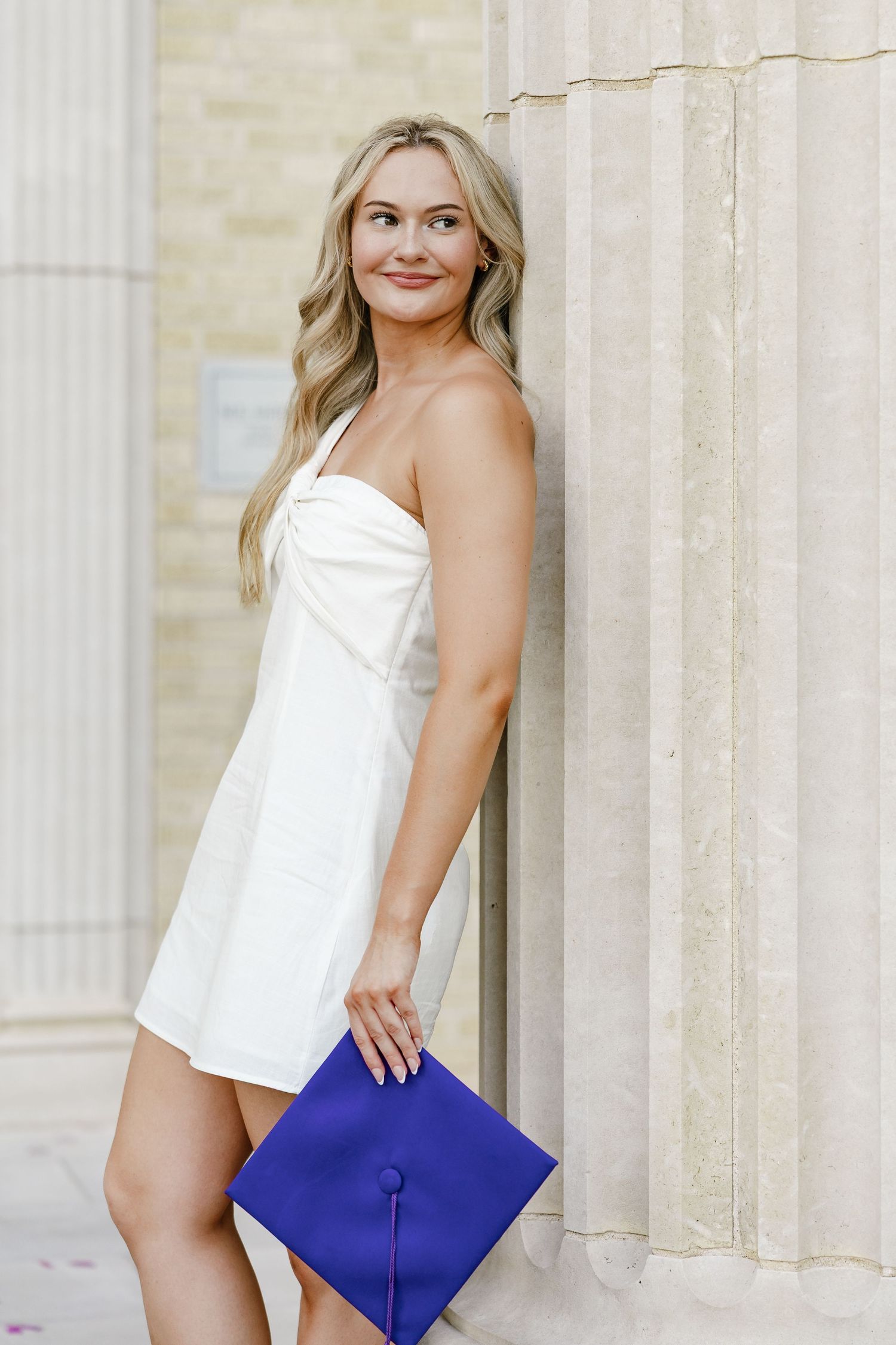 Young blonde woman in white dress holding blue graduation cap, leaning against stone column outdoors.