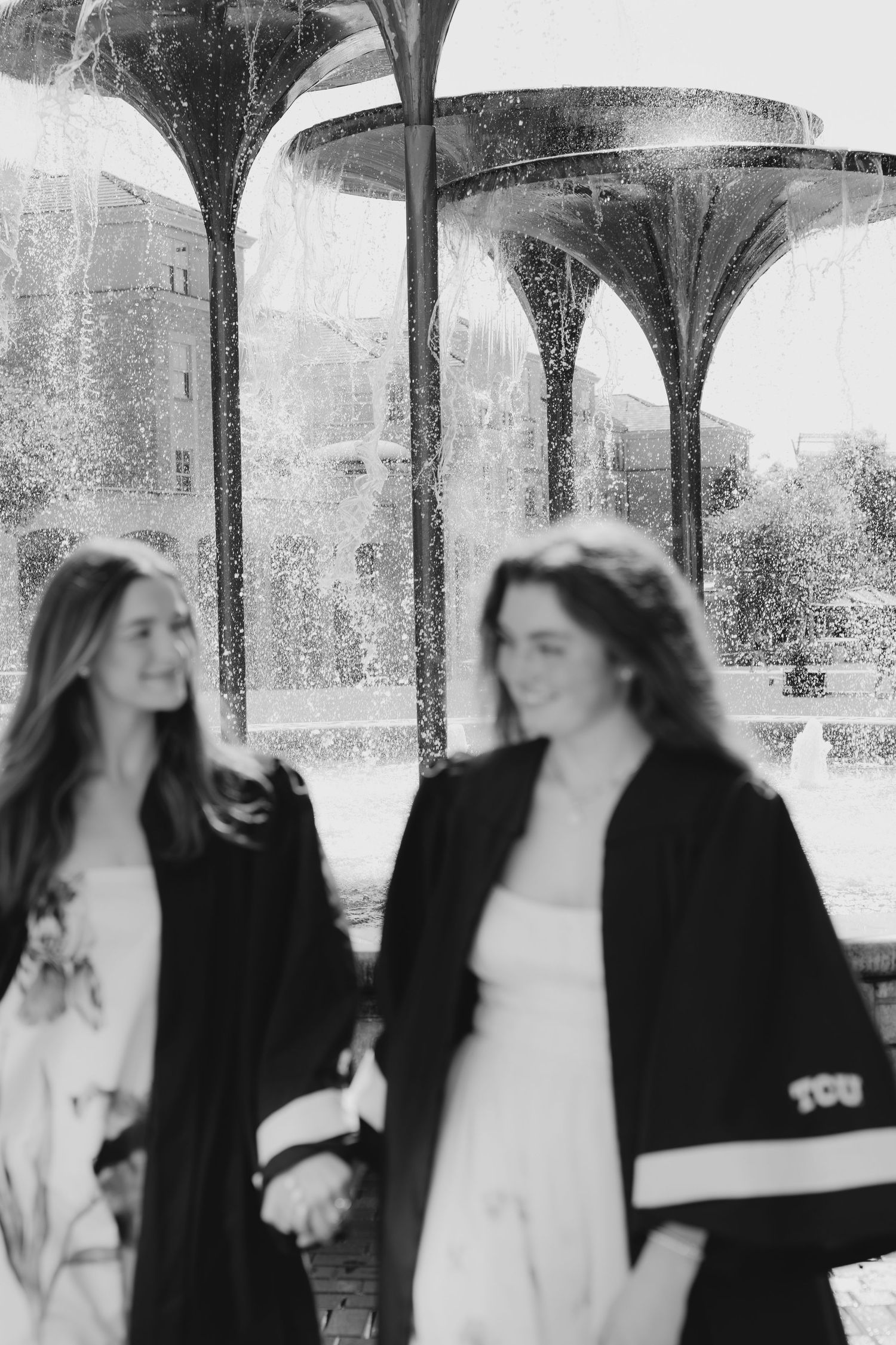 Two young women in graduation gowns hold hands and smile near a fountain in a black and white photo.
