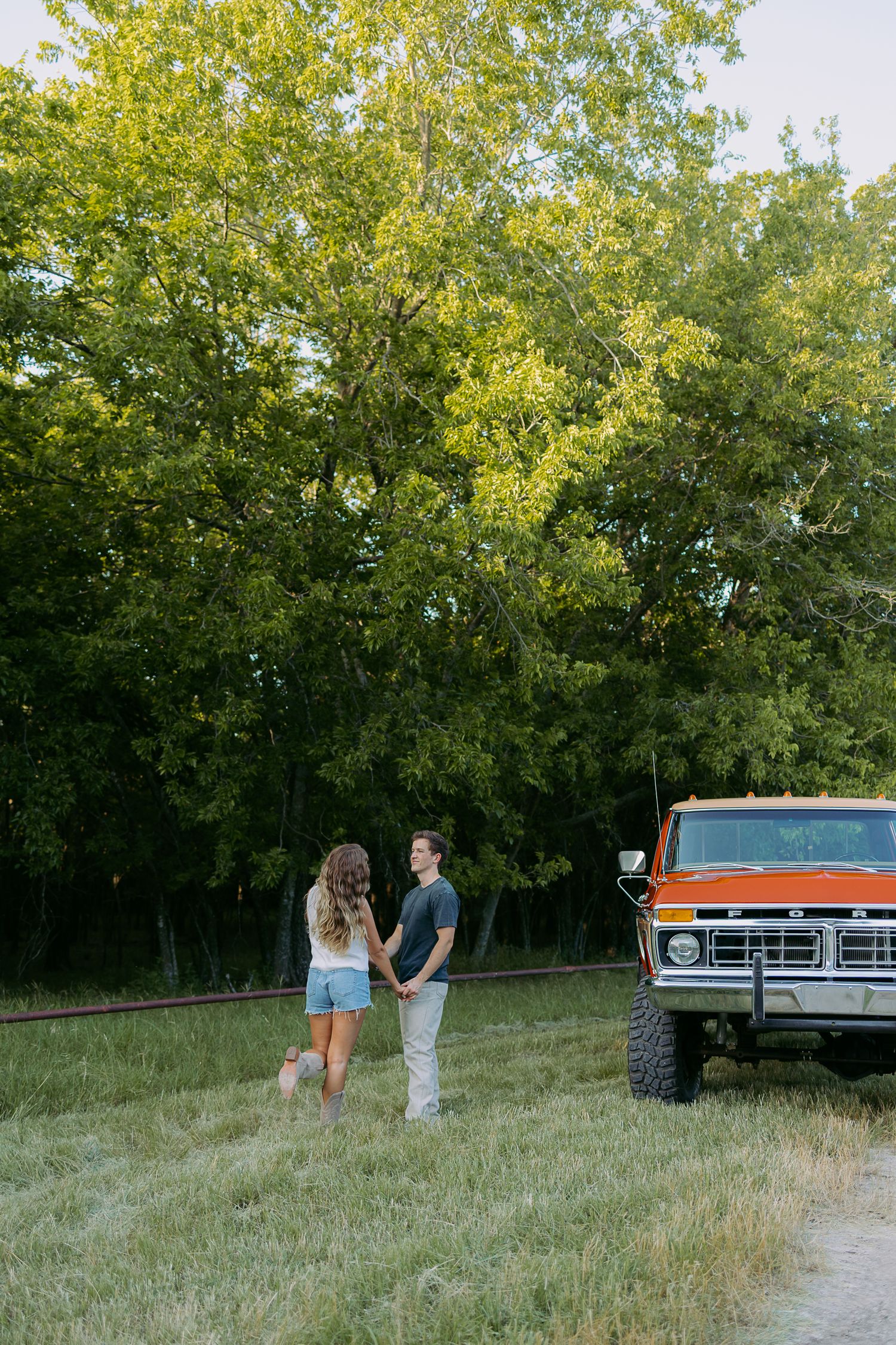 A couple stands near a vintage pickup truck in a grassy field surrounded by trees at golden hour.